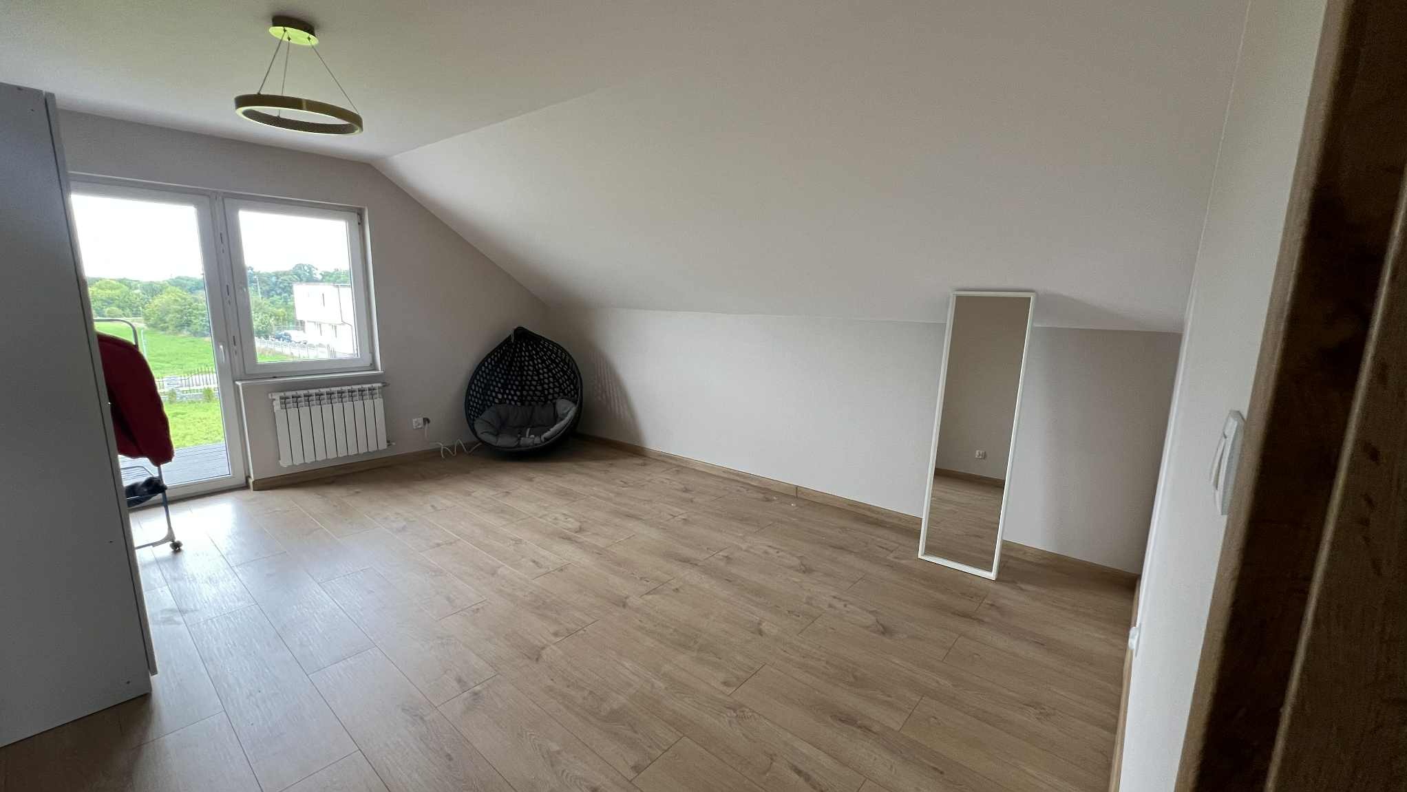 Jasne poddasze po remoncie: drewniana podłoga, białe ściany, okno z widokiem na zieleń. Wnętrze dopełnia wiszący fotel i wysokie lustro w białej ramie.