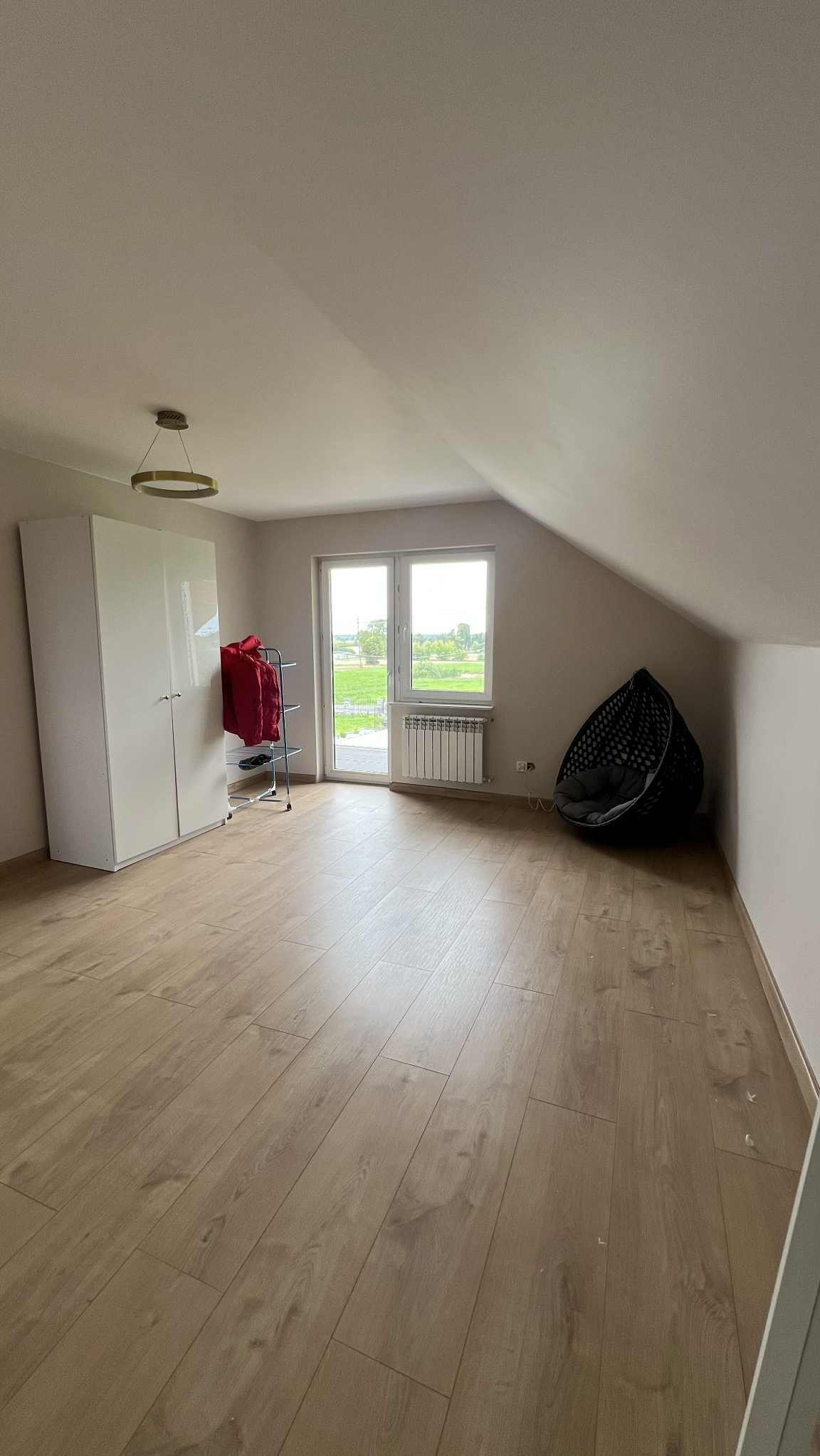 Jasne poddasze z drewnianą podłogą, białymi ścianami i szafą. Widoczne okno z widokiem na zieleń i wiszący fotel. Minimalistyczny design wnętrza.