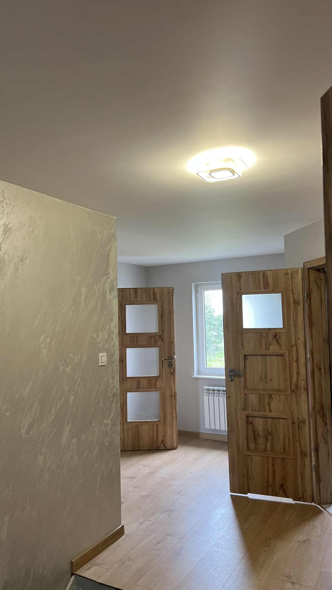 Nowoczesne wnętrze z dwiema drewnianymi drzwiami z przeszkleniami, wykończone ściany z efektem dekoracyjnym i jasna podłoga. Widok na okno z grzejnikiem.