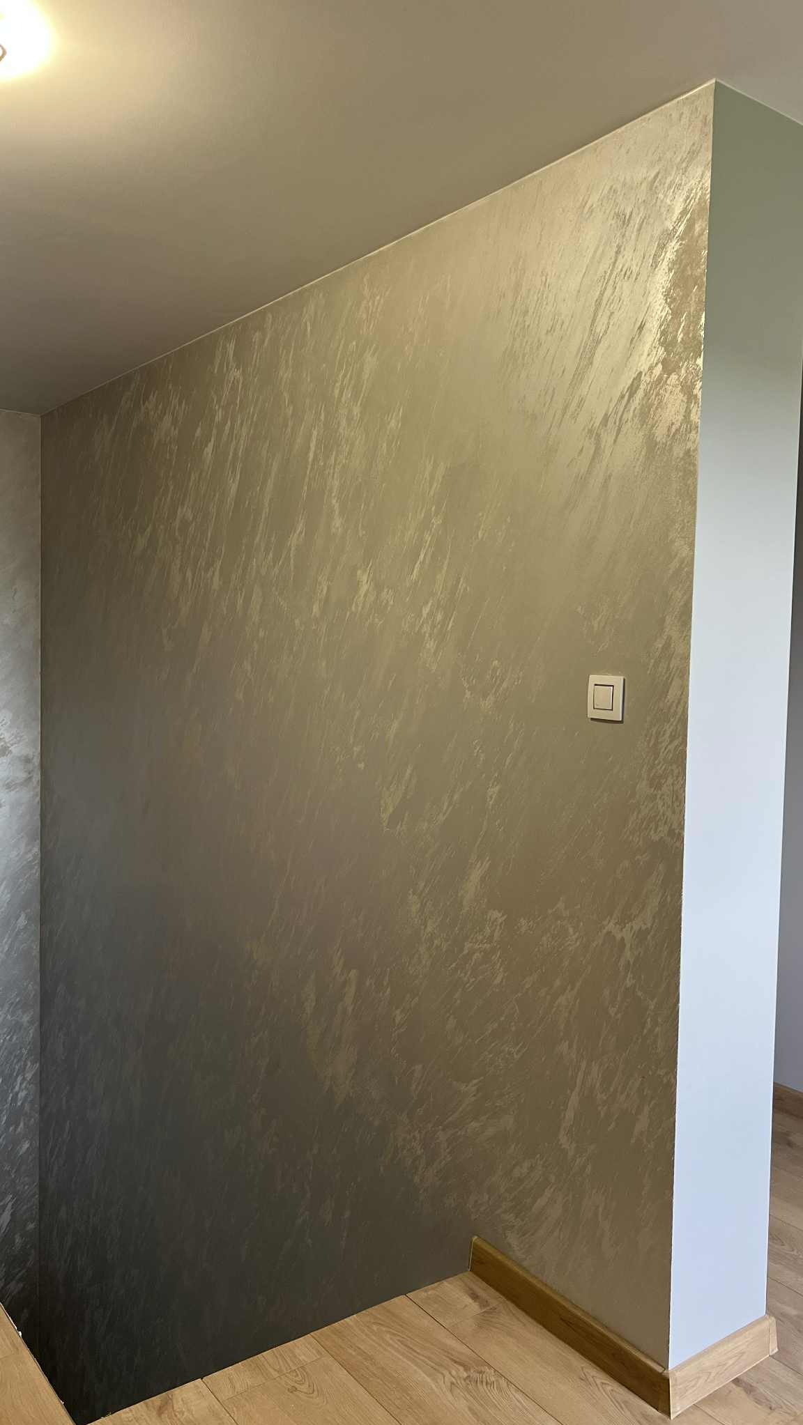 Ściana z dekoracyjnym, srebrzystoszarym efektem malowania, widoczny włącznik światła i fragment podłogi z drewnianą listwą przypodłogową.