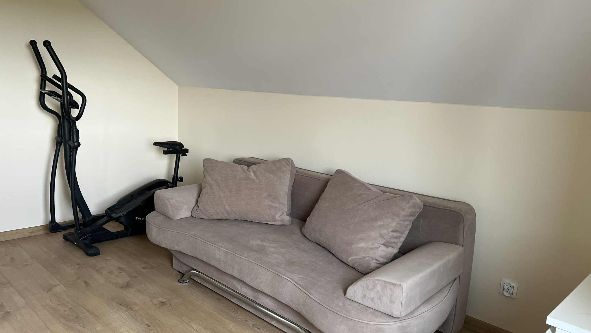 Jasna sofa z poduszkami w pokoju na poddaszu we Włocławku. Obok sprzęt do ćwiczeń. Neutralna kolorystyka ścian i podłogi. Widok na wnętrze pokoju.