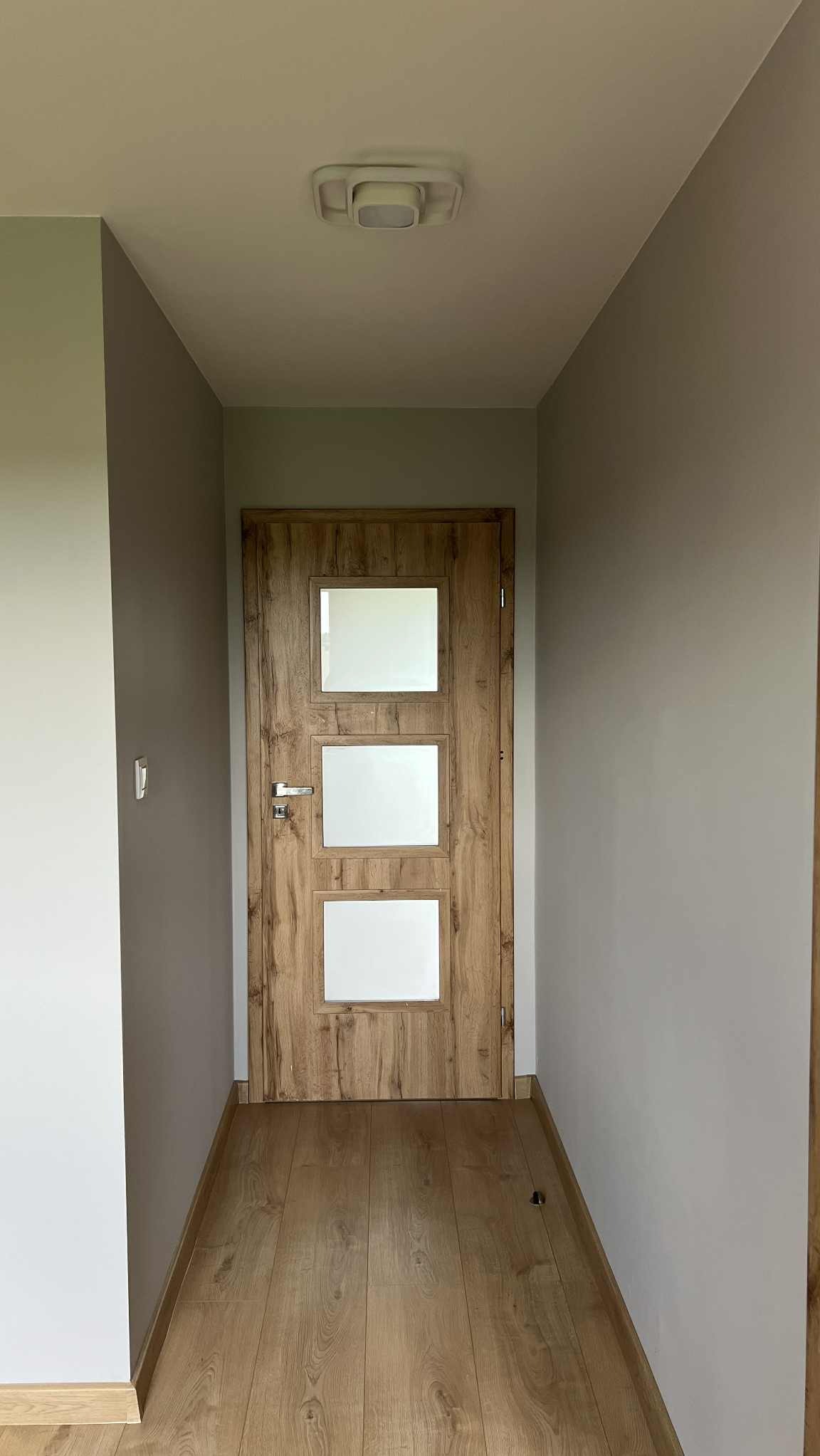 Drewniane drzwi wewnętrzne z trzema szybami w odcieniach bieli, zamontowane w szarej ścianie, widoczne z perspektywy korytarza z panelami podłogowymi.