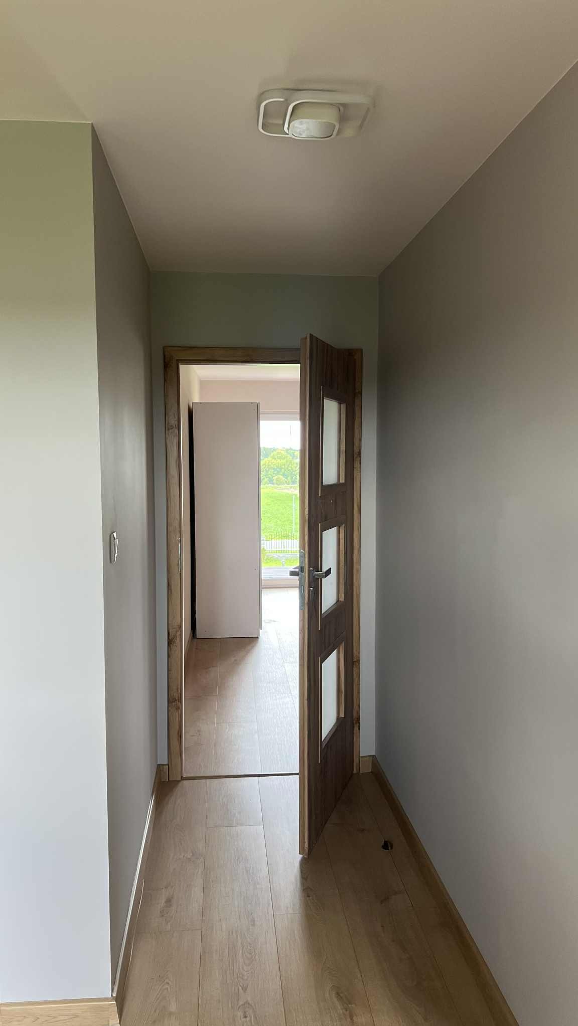 Korytarz z drewnianą podłogą i drzwiami wewnętrznymi z przeszkleniami, pomalowany na szaro. Widok na kolejne pomieszczenie z oknem i zielenią za nim.