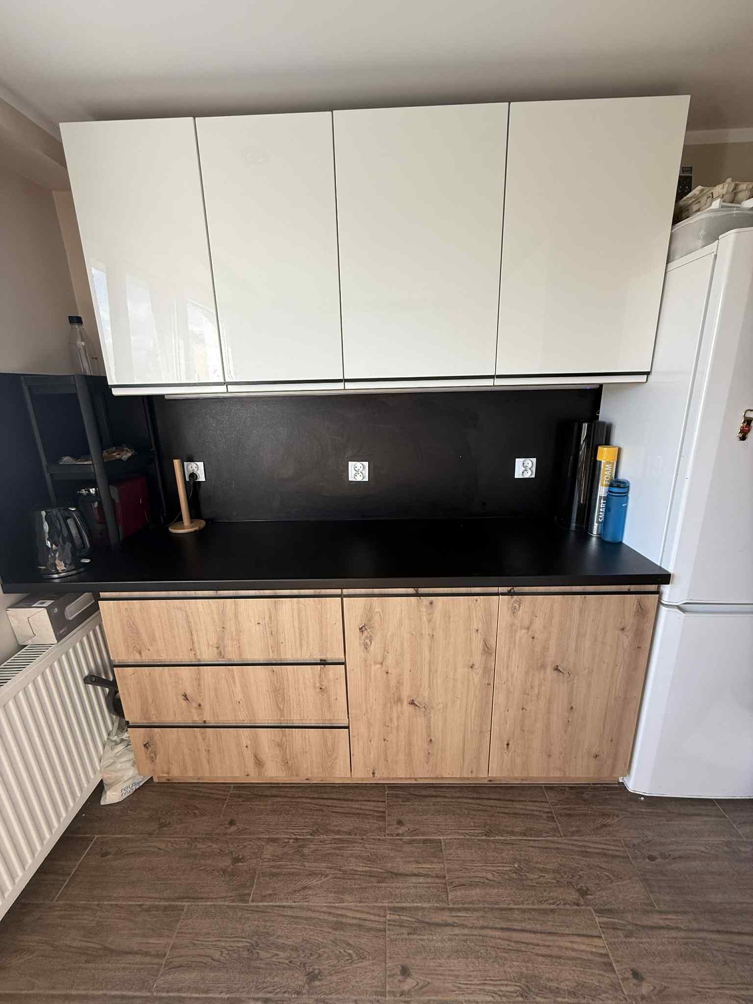 Nowoczesna zabudowa kuchenna: białe szafki górne, drewnopodobne dolne z czarnym blatem i ścianą, widoczne gniazdka elektryczne. Ukończony remont kuchni.