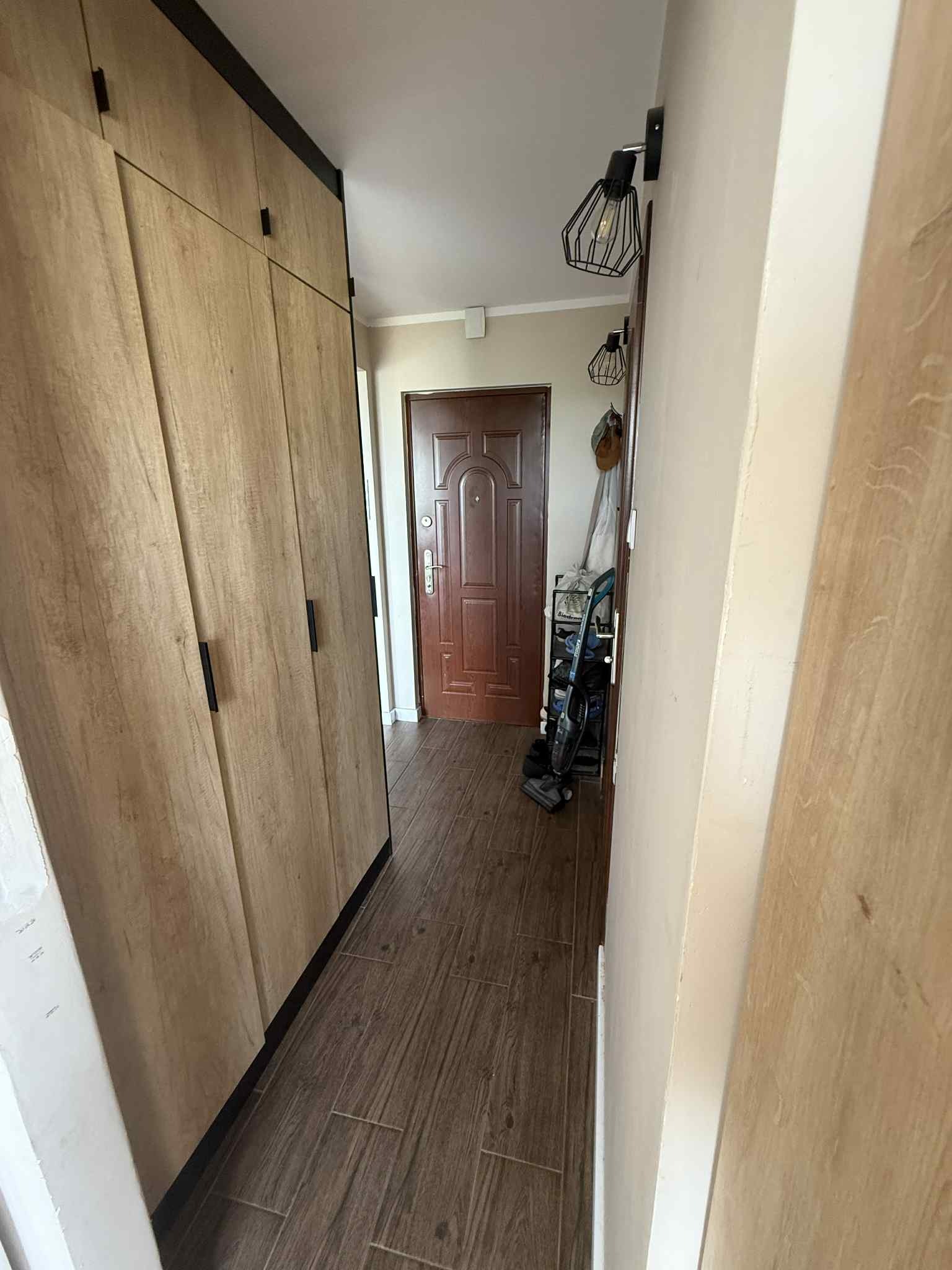 Wnętrze przedpokoju z drewnianą podłogą i zabudowaną szafą z drewnopodobnymi frontami. Widoczne czarne, geometryczne lampy sufitowe i brązowe drzwi wejściowe.