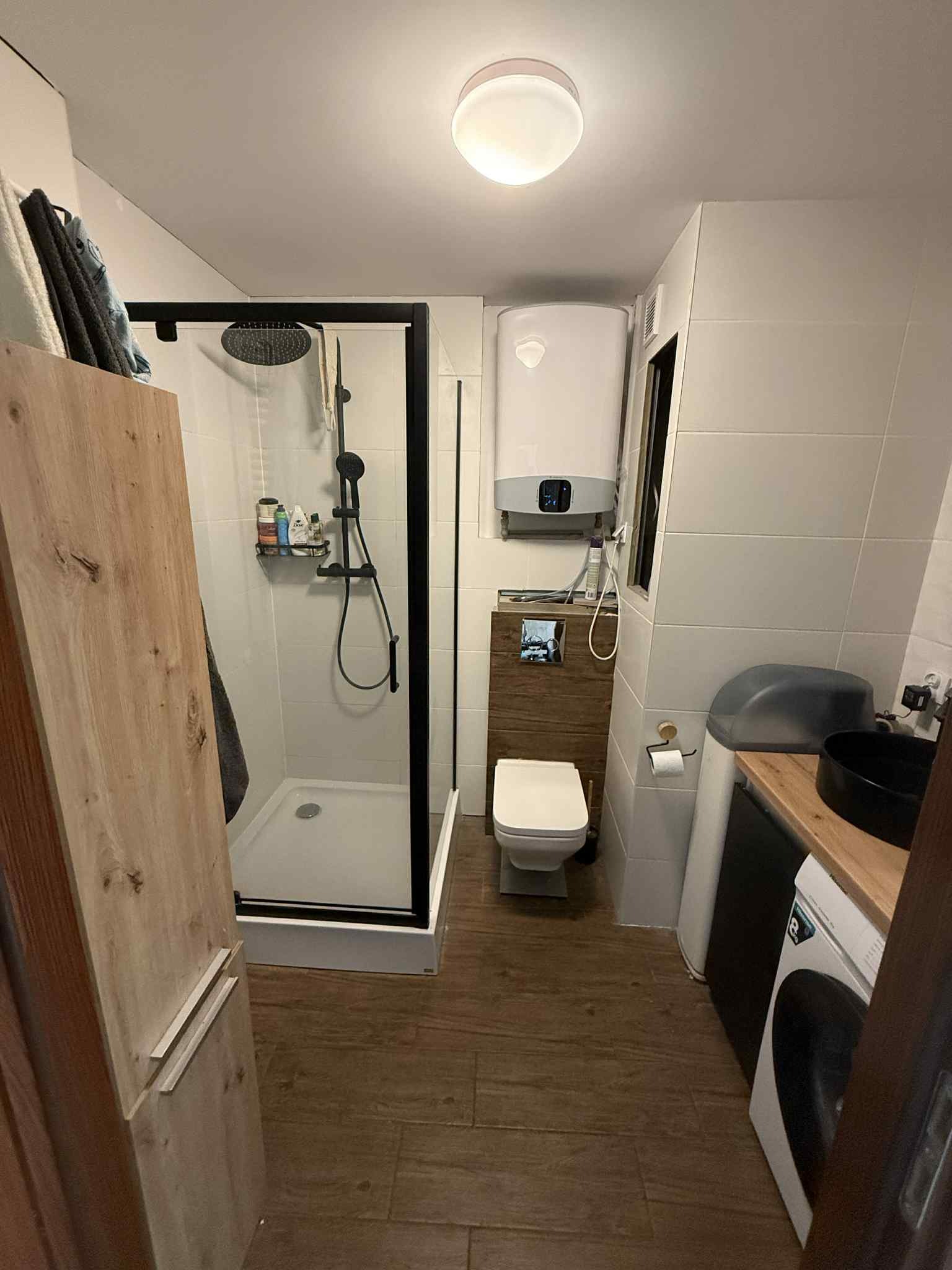 Nowoczesna łazienka z kabiną prysznicową typu walk-in, wiszącą toaletą, drewnianymi akcentami i białymi płytkami. Widoczny bojler i pralka.