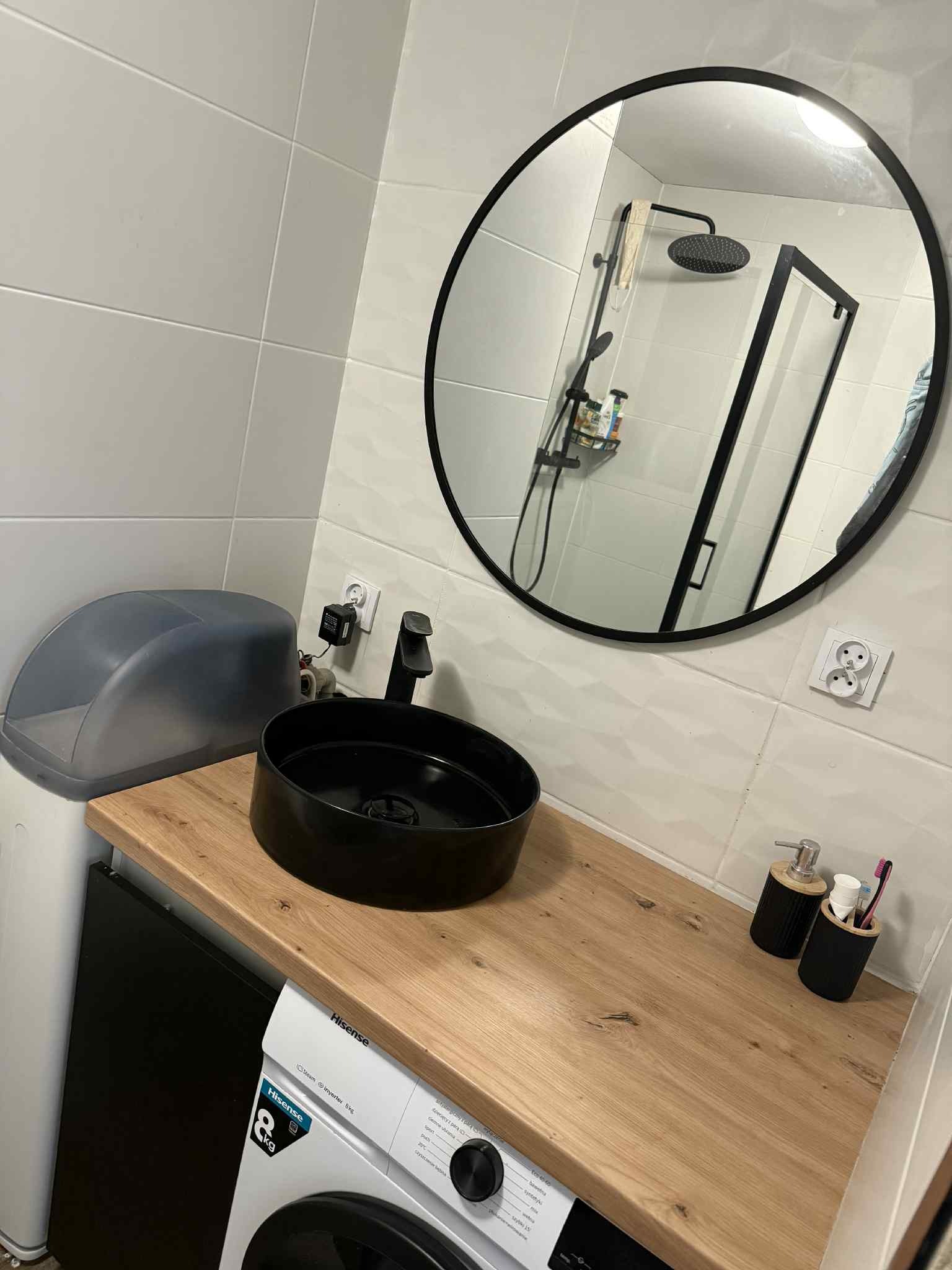 Nowoczesna łazienka z czarną umywalką na drewnianym blacie, okrągłe lustro odbija kabinę prysznicową. Minimalistyczny design, białe płytki.