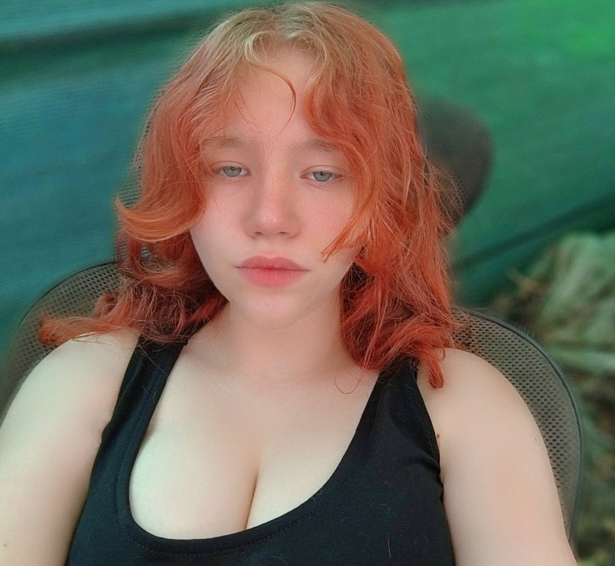 Portret młodej kobiety z rudymi, kręconymi włosami i jasną cerą, ubrana w czarny top na tle zieleni. Ujęcie z bliska, skupione na twarzy i włosach.