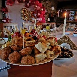 Estera Kołodziej - Uroczysty stół z przekąskami, balonami w kształcie cyfr 85 w tle, świeca. Catering na imprezę okolicznościową z dekoracjami.