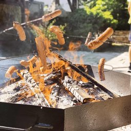 Estera Kołodziej - Ognisko w metalowej skrzyni z unoszącymi się nad nim kiełbaskami nabitymi na patyki. W tle osoby przygotowujące posiłek na świeżym powietrzu.