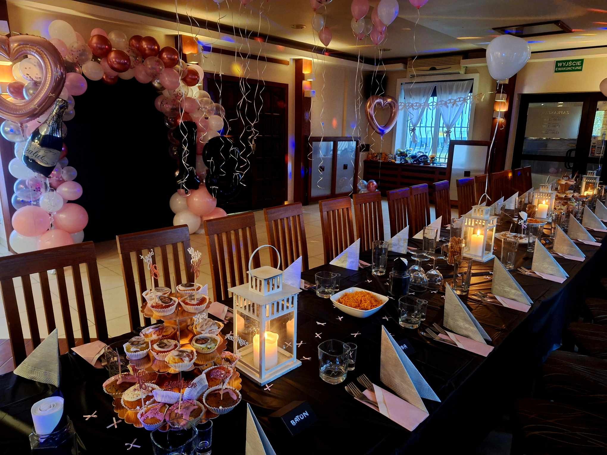 Uroczysty stół z czarnym obrusem, dekoracjami balonowymi, babeczkami i lampionami, przygotowany na przyjęcie. Widoczne nakrycia i dekoracje.