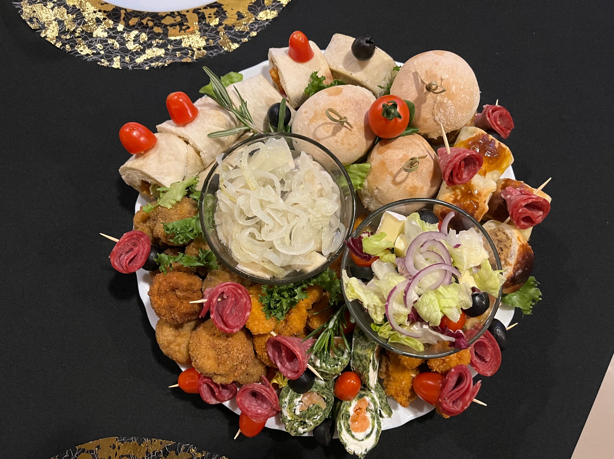 Dekoracyjna kompozycja cateringowa na białym talerzu: mini-kanapki, roladki ze szpinakiem i łososiem, sałatki w szklanych miseczkach, koreczki z salami i pomidorkami koktajlowymi.