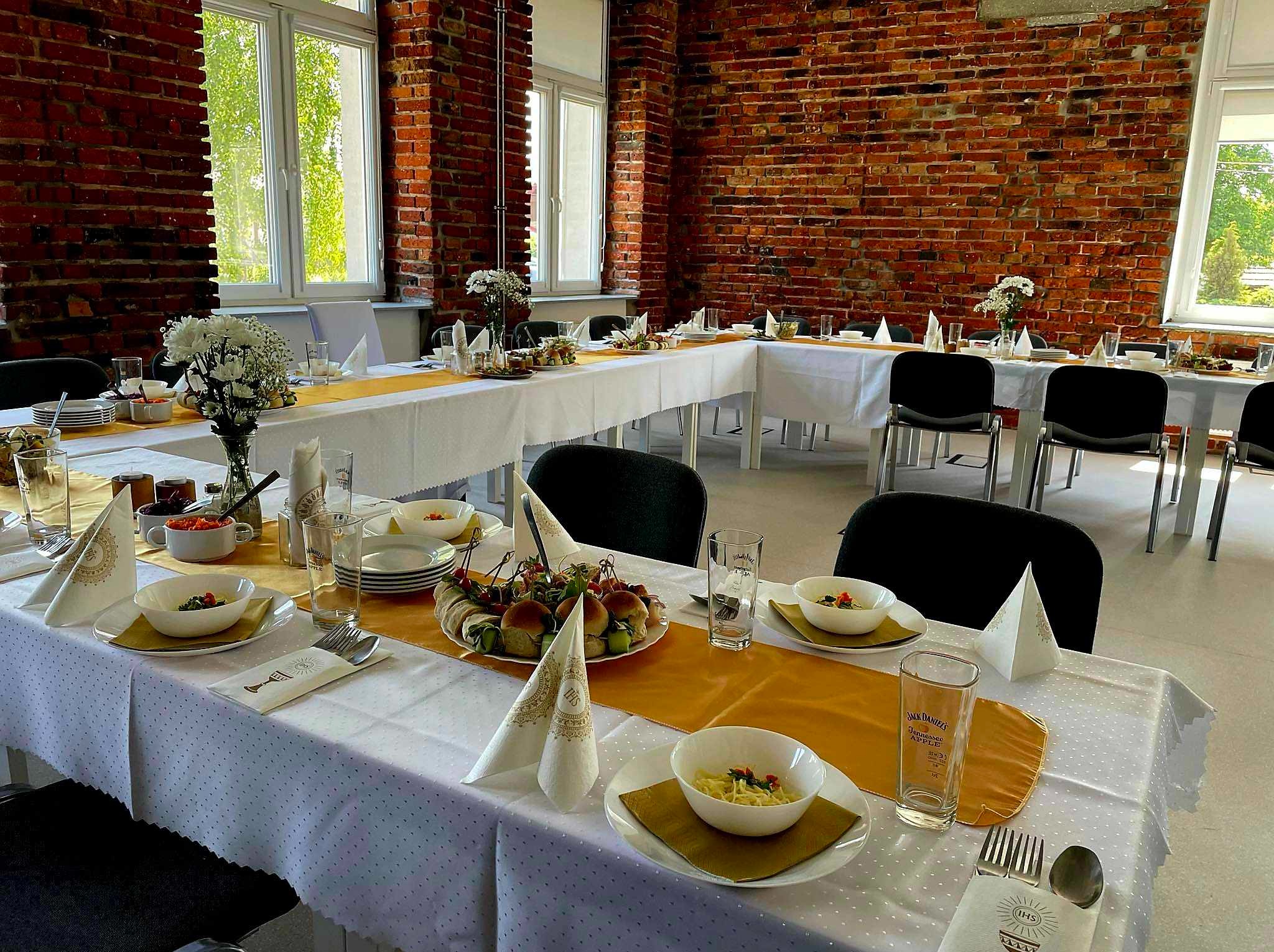 Uroczysty stół z białym obrusem, złotym bieżnikiem, zastawą i dekoracjami kwiatowymi na tle ceglanej ściany i okien. Przygotowany na przyjęcie.