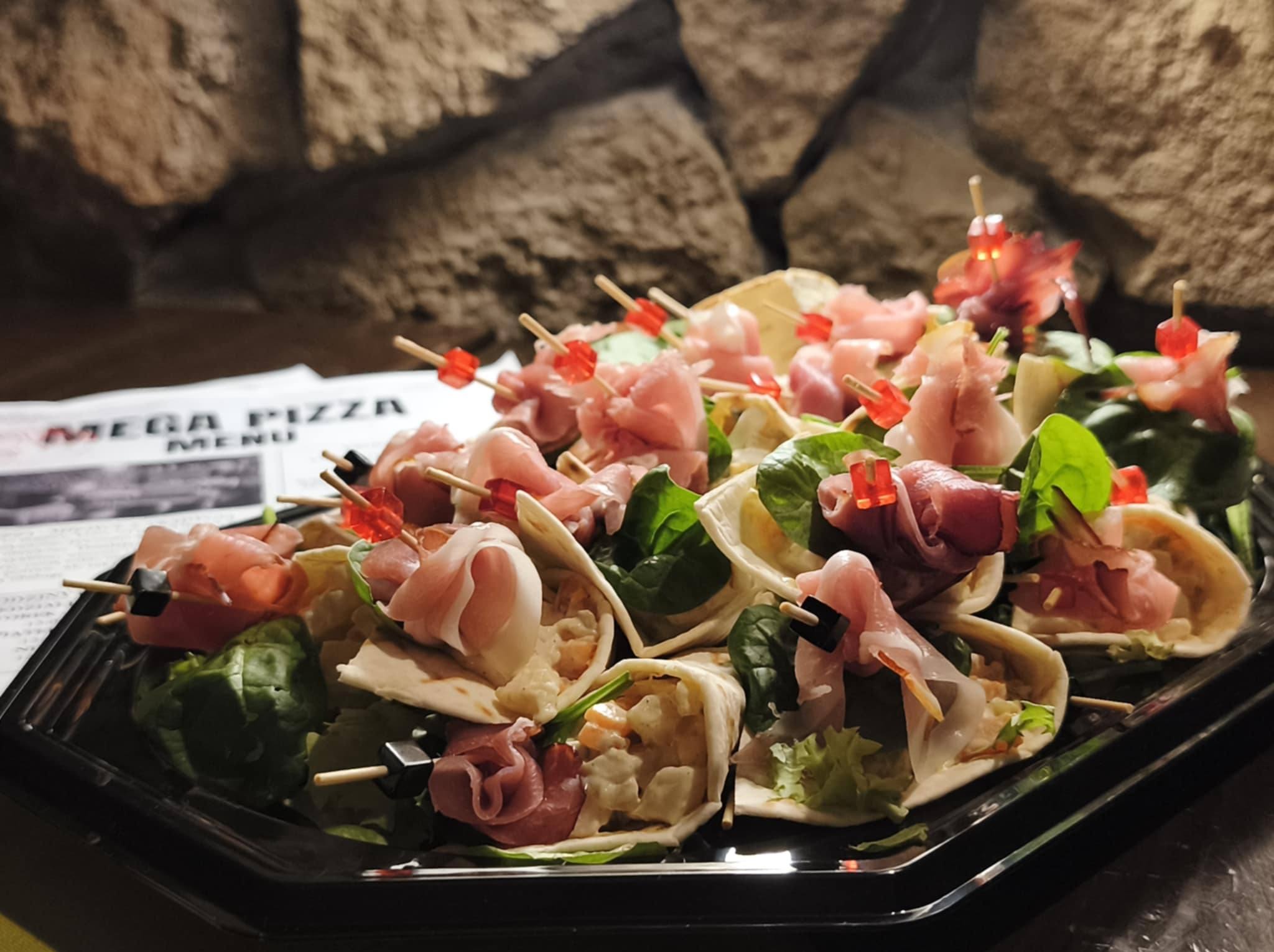 Elegancki catering: tortille z szynką parmeńską i szpinakiem na czarnej tacy, dekorowane kolorowymi wykałaczkami, na tle kamiennej ściany i menu Mega Pizza.