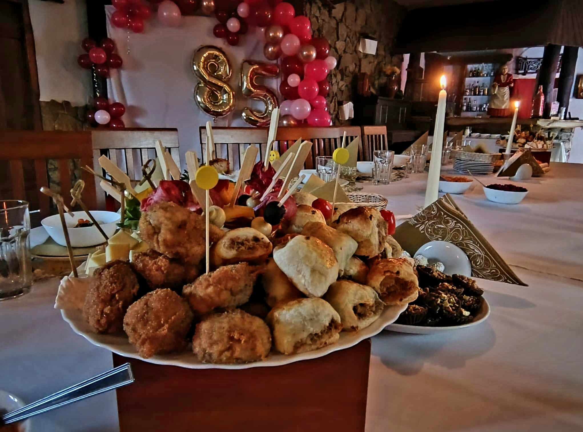 Uroczysty stół z przekąskami, balonami w kształcie cyfr 85 w tle, świeca. Catering na imprezę okolicznościową z dekoracjami.