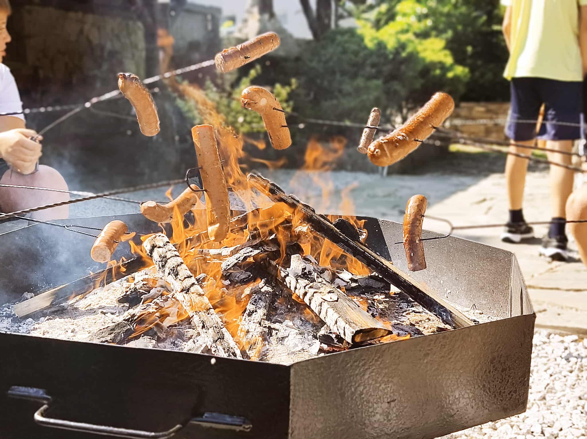 Ognisko w metalowej skrzyni z unoszącymi się nad nim kiełbaskami nabitymi na patyki. W tle osoby przygotowujące posiłek na świeżym powietrzu.