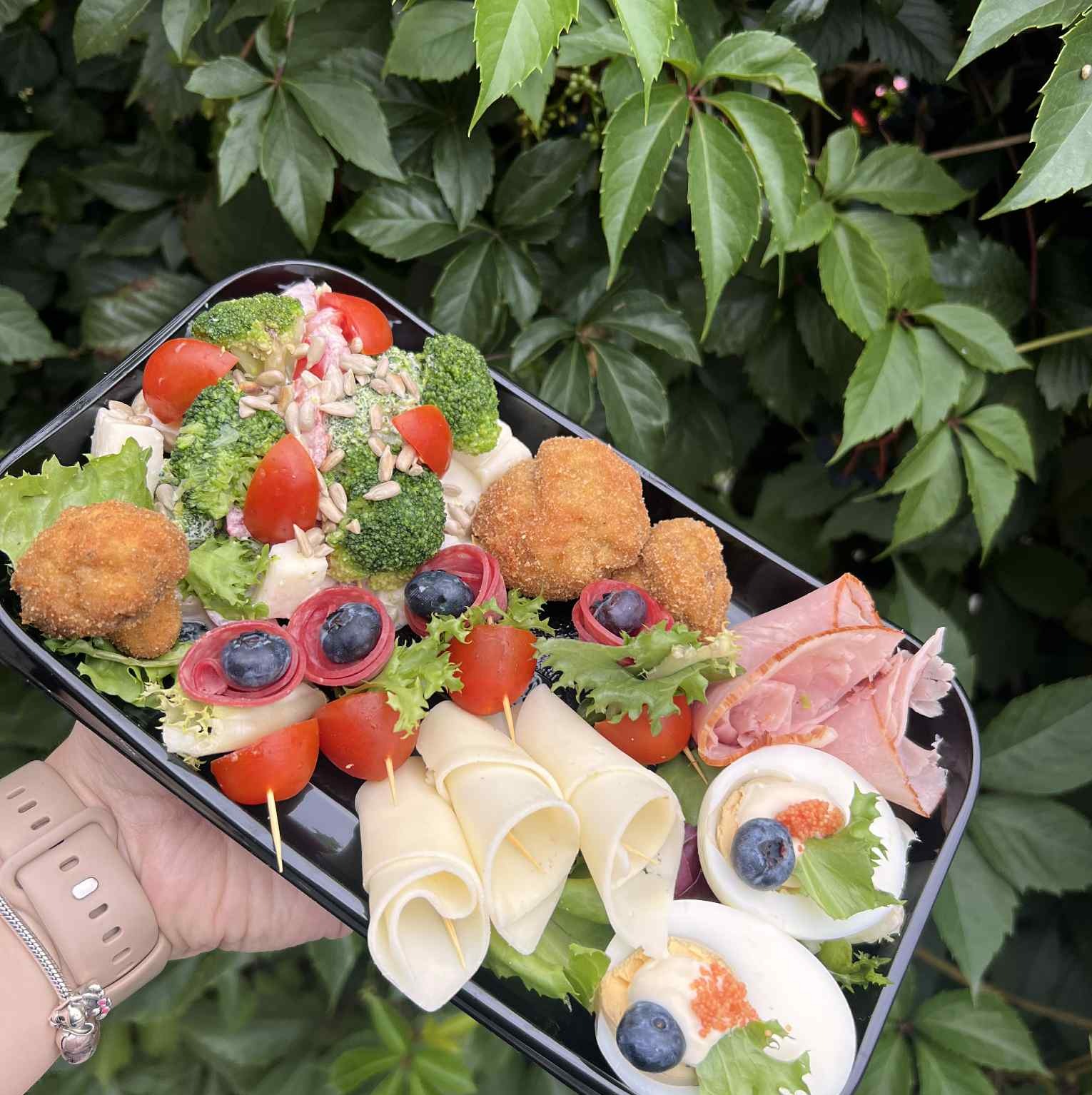 Czarny talerz z kompozycją przekąsek: sałatka z brokułami, jajka z kawiorem i borówkami, roladki z sera i szynki, kulki serowe, pomidorki koktajlowe na tle zieleni.