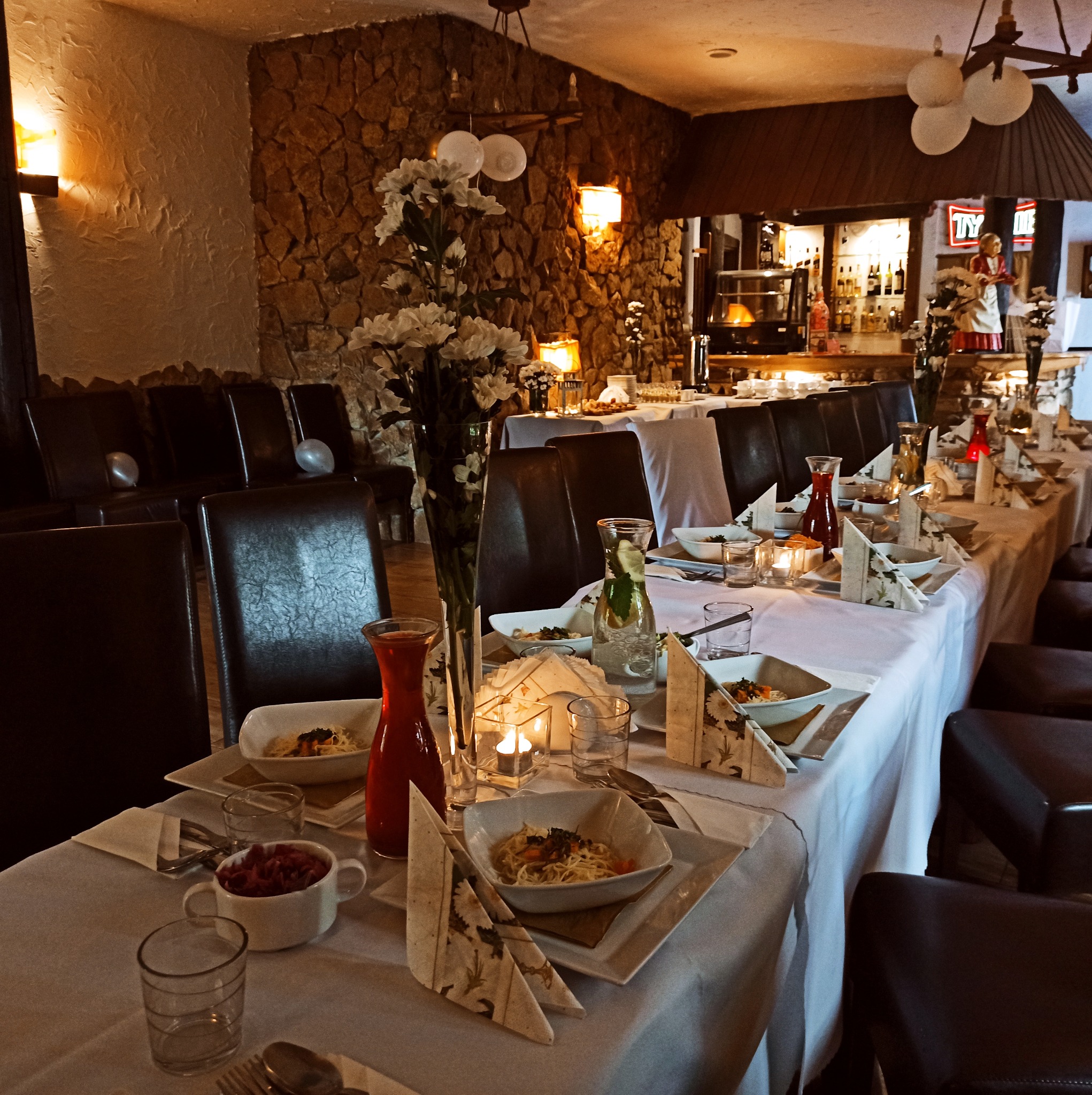 Uroczysty stół z białym obrusem, zastawą i dekoracjami kwiatowymi w restauracji o kamiennych ścianach, przygotowany na przyjęcie.