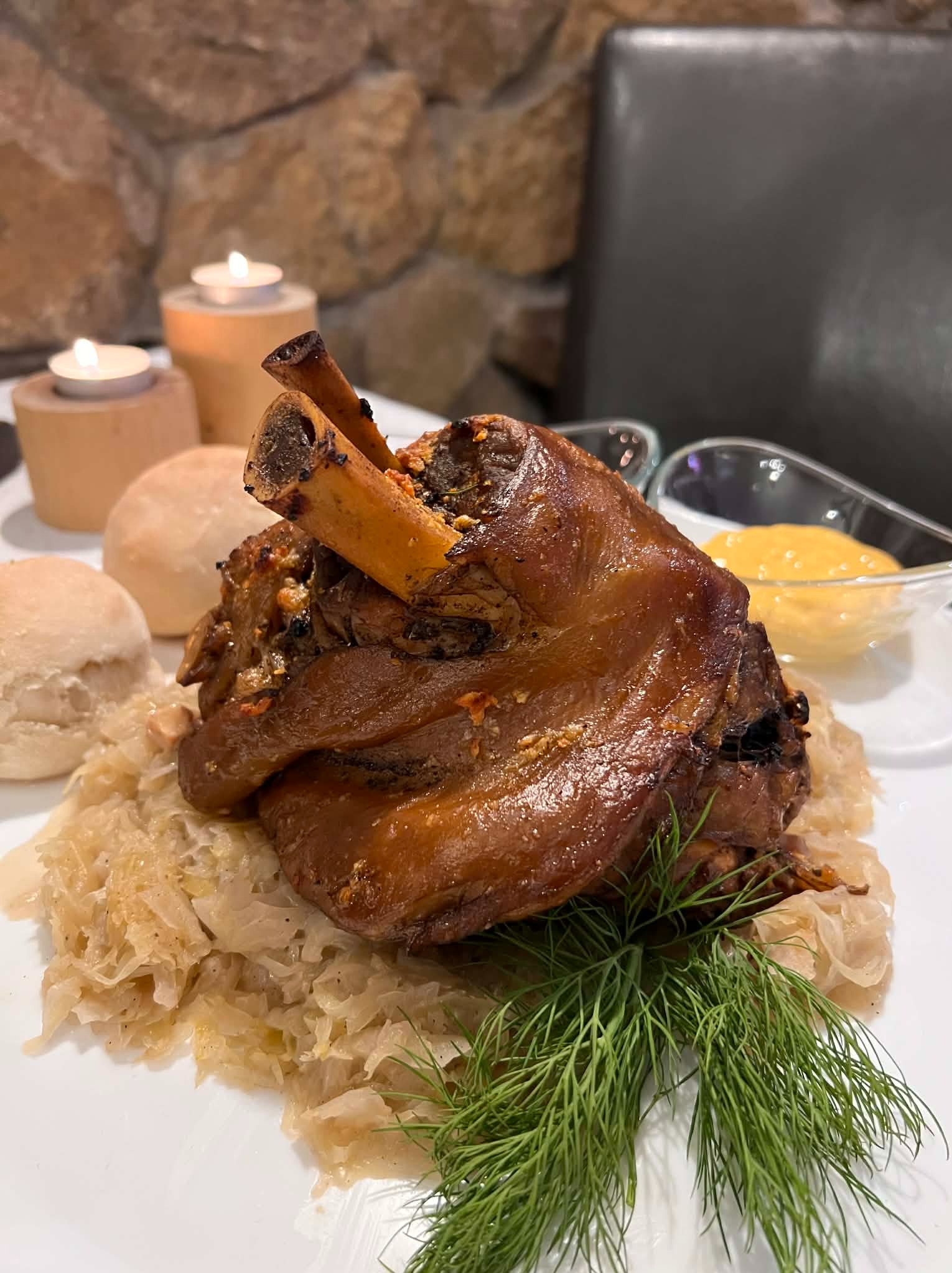 Apetyczna golonka na kapuście, z dodatkiem świeżego koperku, bułeczek i sosu, podana na białym talerzu. W tle dekoracyjne świece i kamienna ściana.