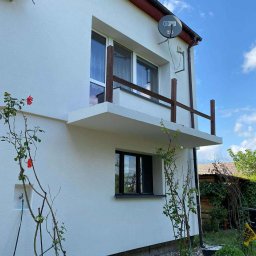 SMARTTHERM SPÓŁKA Z OGRANICZONĄ ODPOWIEDZIALNOŚCIĄ - Biały dom z balkonem, klimatyzatorem Hawaii i anteną satelitarną. Widoczne róże przy ścianie i fragment ogrodu. Kadr z dołu, niebo z chmurami w tle.