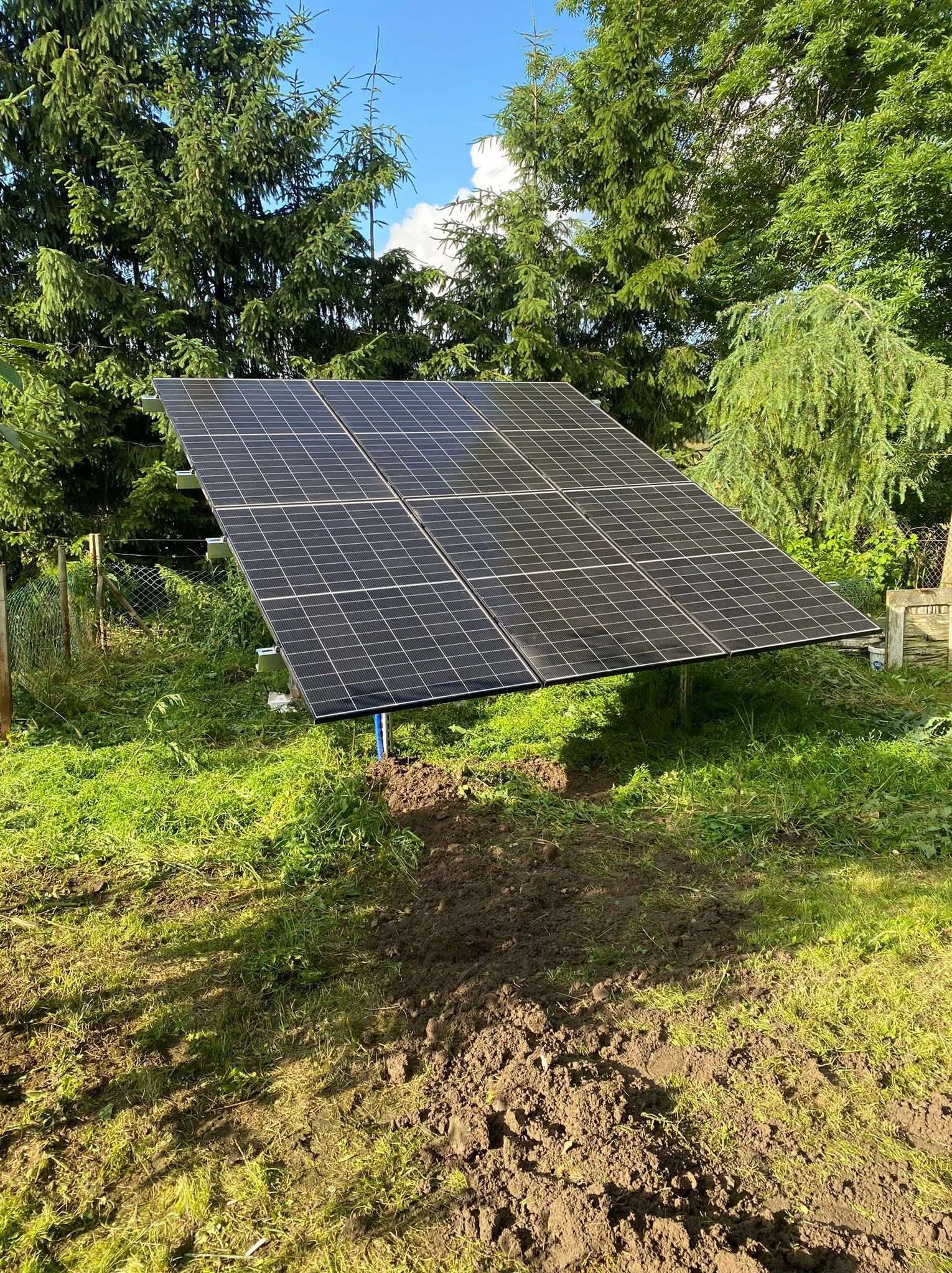 Ustawiony panel fotowoltaiczny na trawniku, widok z dołu. Cztery czarne panele na metalowej konstrukcji, tło z zieleni drzew i błękitnego nieba.