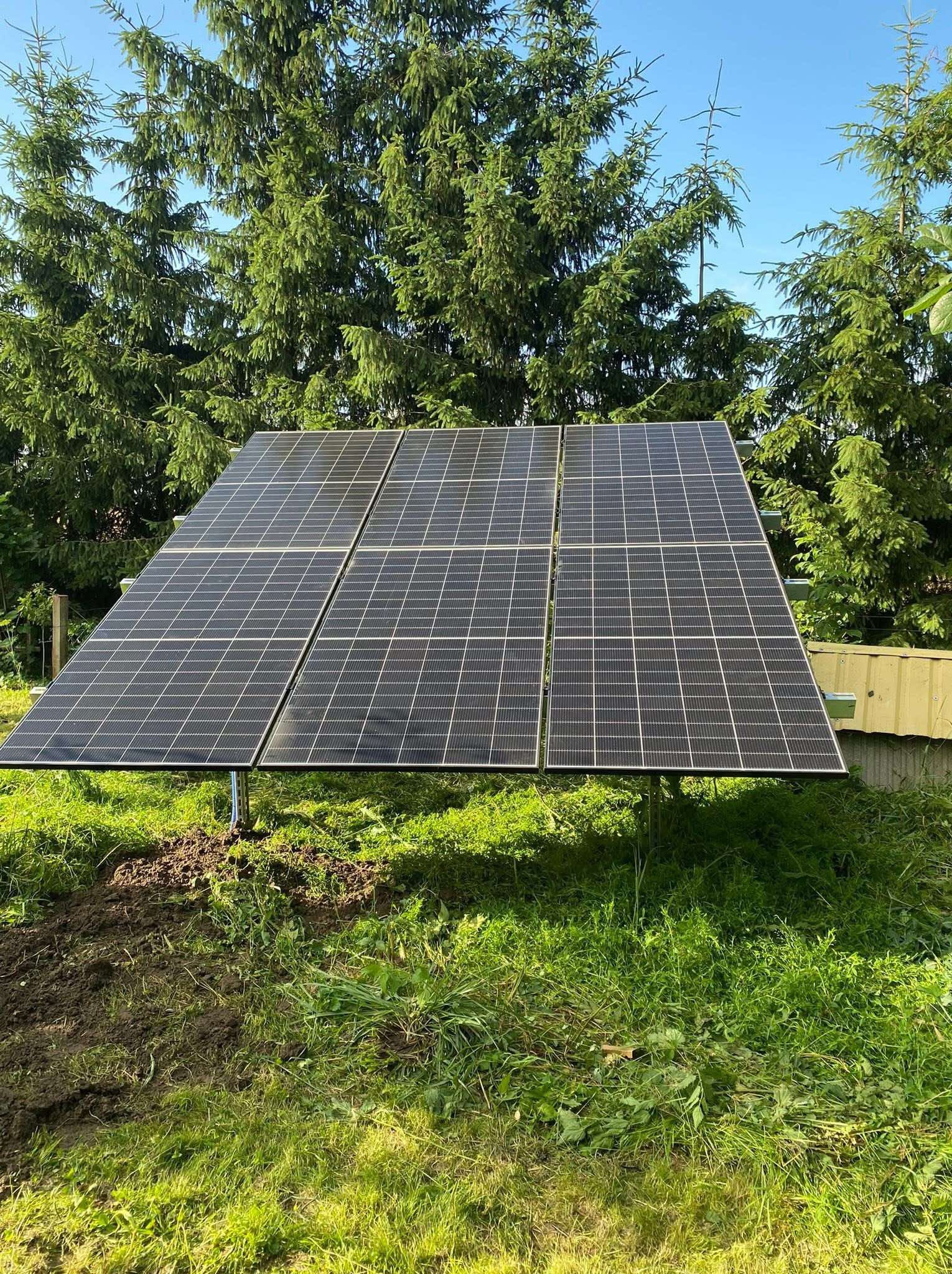 Cztery panele fotowoltaiczne zamontowane na metalowej konstrukcji, otoczone zielenią i drzewami w słoneczny dzień. Widoczne kable i przygotowane podłoże.