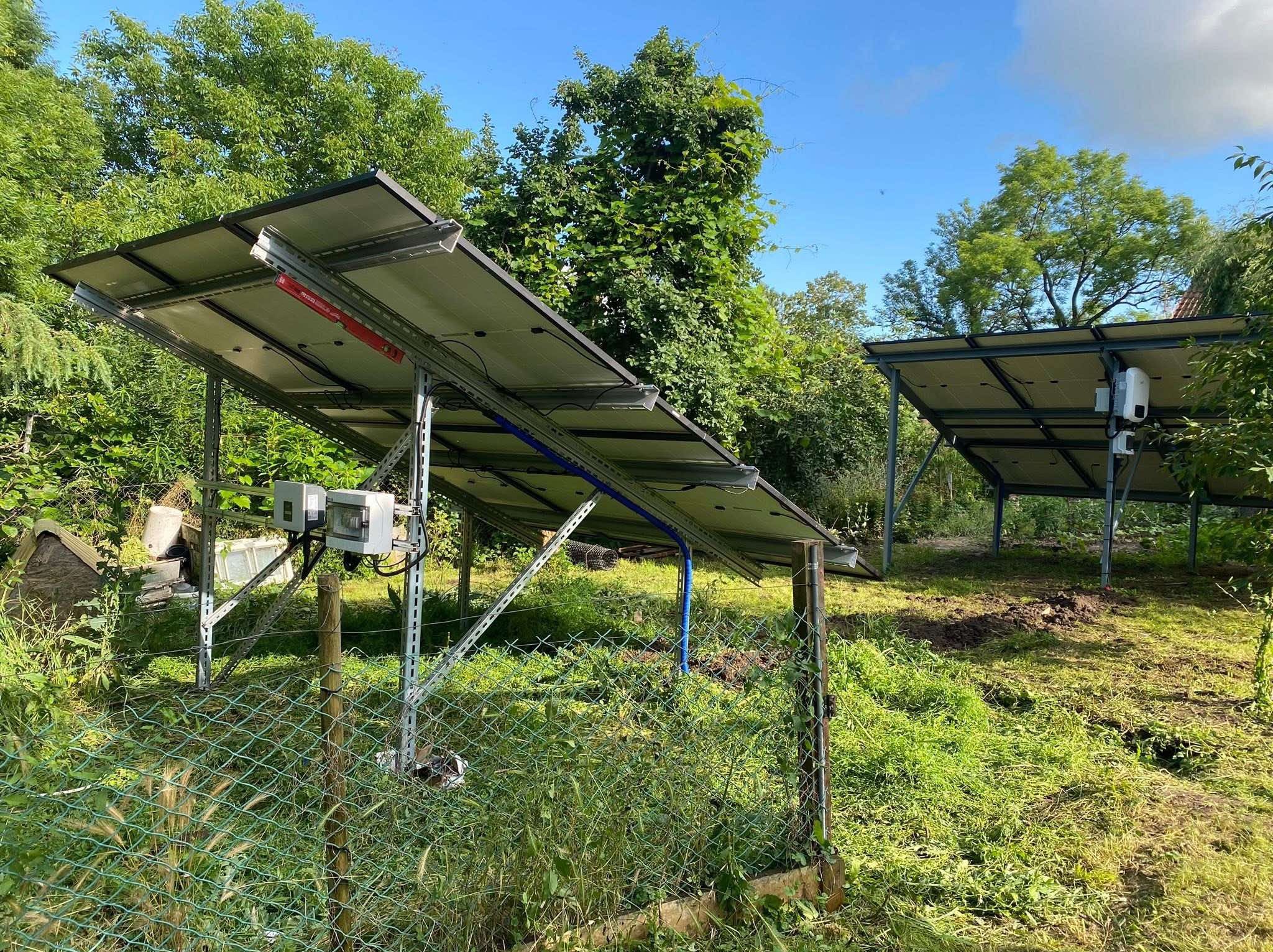Dwie instalacje fotowoltaiczne na metalowych stelażach, widoczne zza zielonego płotu w ogrodzie, z gęstą zielenią w tle i błękitnym niebem.