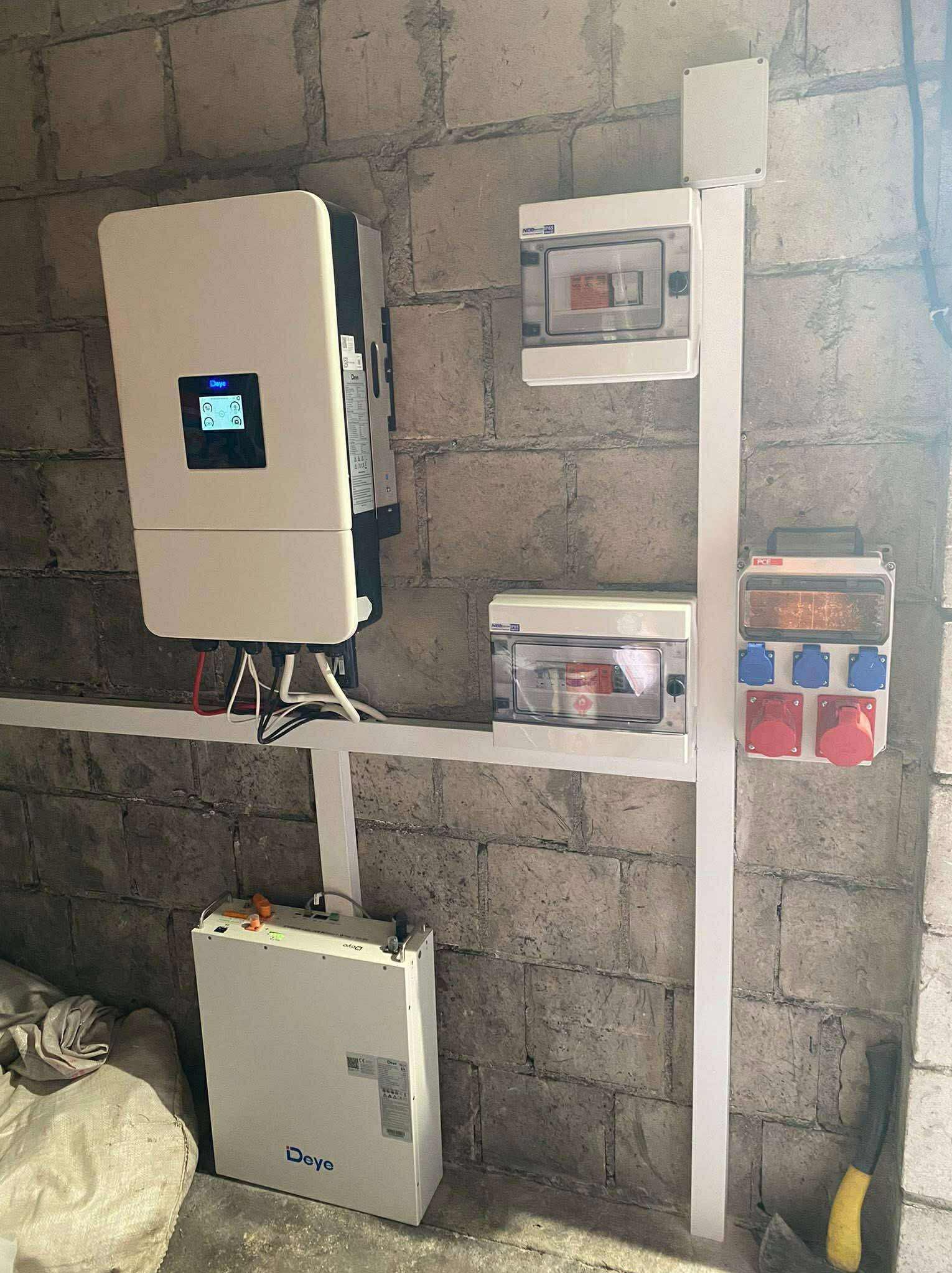 Zamontowany inwerter Deye, akumulator i rozdzielnice elektryczne na szarej, surowej ścianie z cegieł. Białe korytka kablowe. Instalacja fotowoltaiczna wewnątrz budynku.