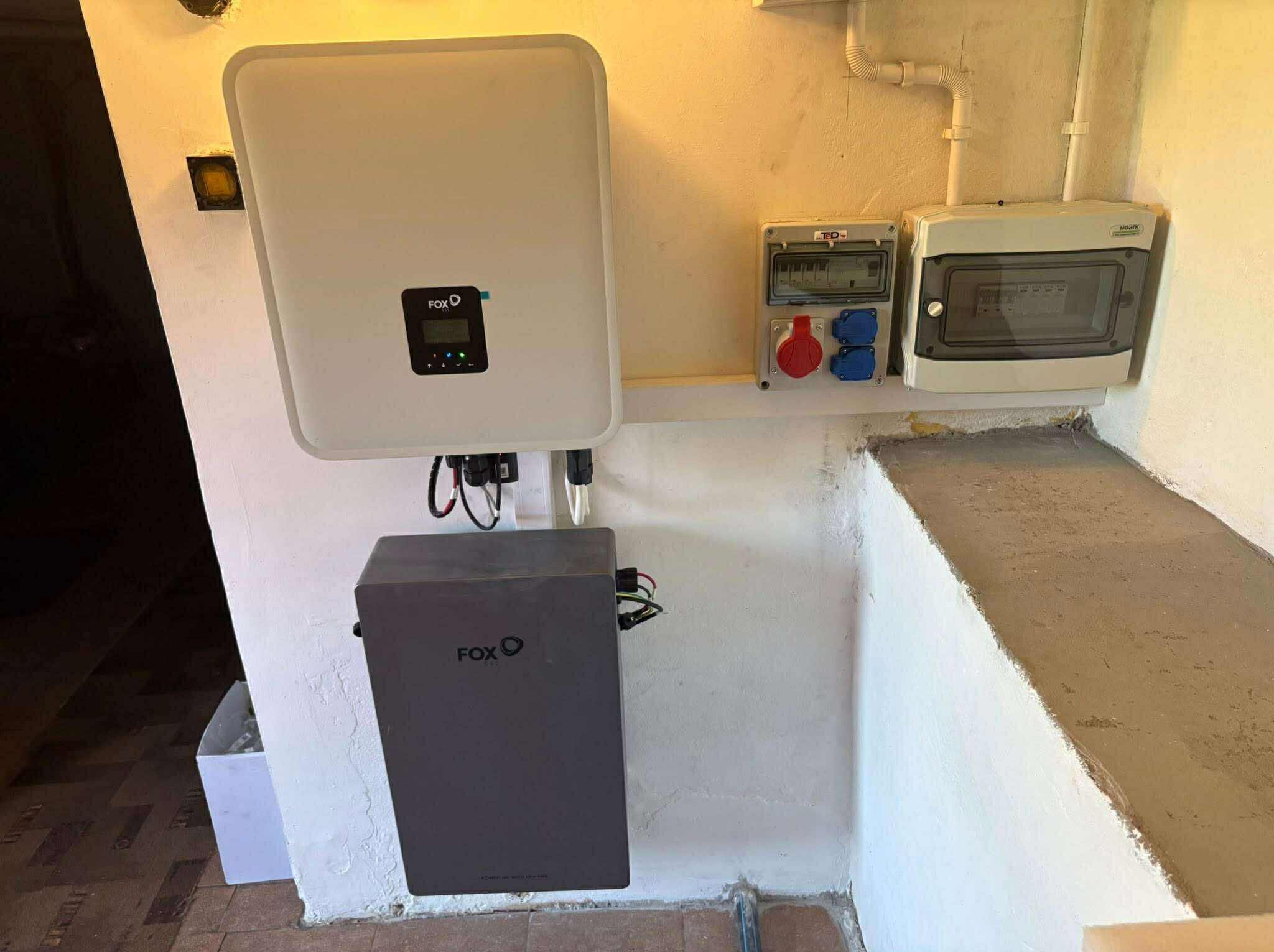 Biały falownik FoxESS z wyświetlaczem i szary akumulator, zamontowane na kremowej ścianie obok skrzynki elektrycznej z gniazdami. Widoczne przewody i złącza.