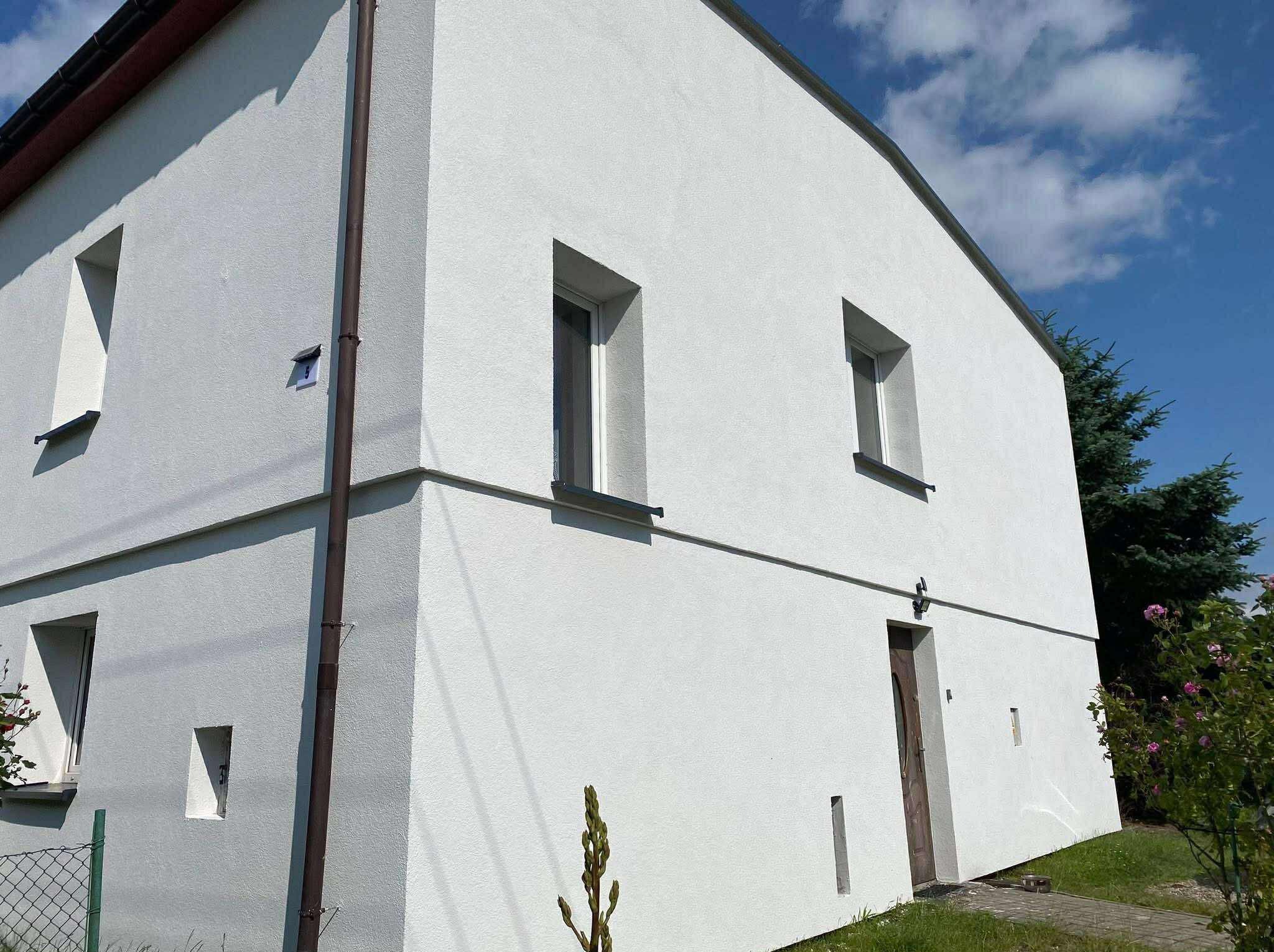 Biała fasada dwukondygnacyjnego domu z brązową rynną, oknami i drzwiami. Widoczne elementy ogrodu i błękitne niebo.