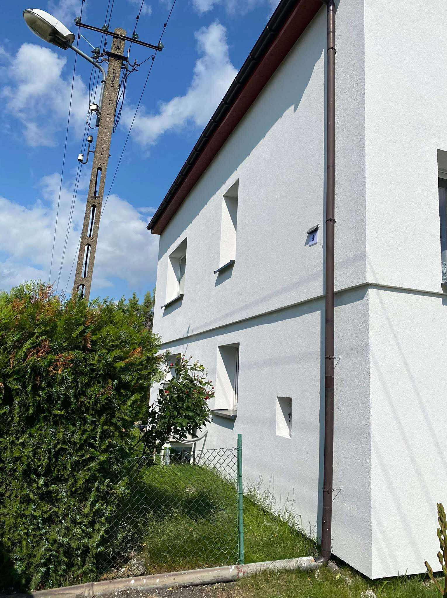 Biały dom dwukondygnacyjny z brązową rynną i numerem 5, widziany z boku, z zielonym żywopłotem i słupem energetycznym na tle błękitnego nieba.