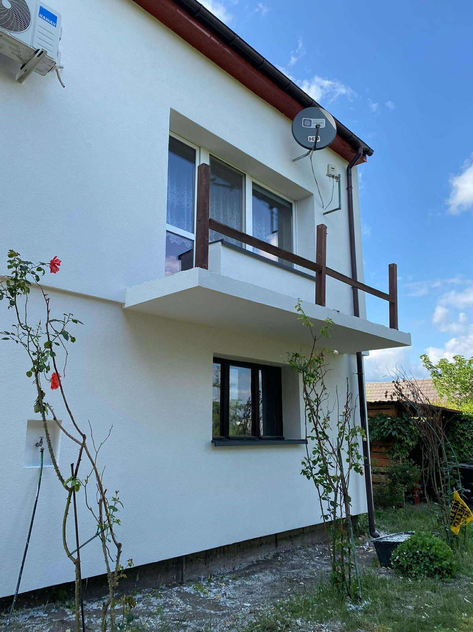 Biały dom z balkonem, klimatyzatorem Hawaii i anteną satelitarną. Widoczne róże przy ścianie i fragment ogrodu. Kadr z dołu, niebo z chmurami w tle.