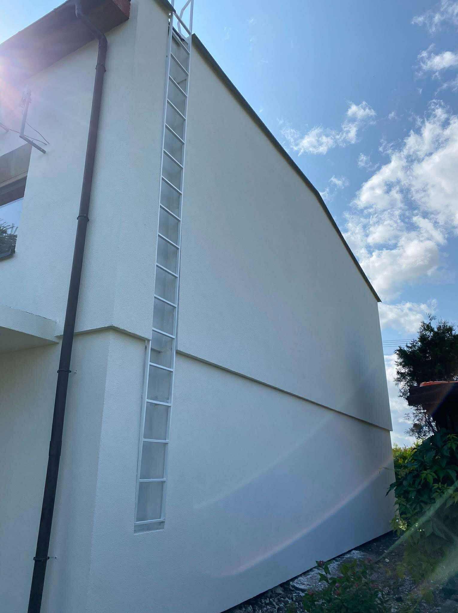 Biała ściana budynku z zamontowaną, pionową drabinką aluminiową. Widoczny fragment rynny. Błękitne niebo z chmurami w tle.