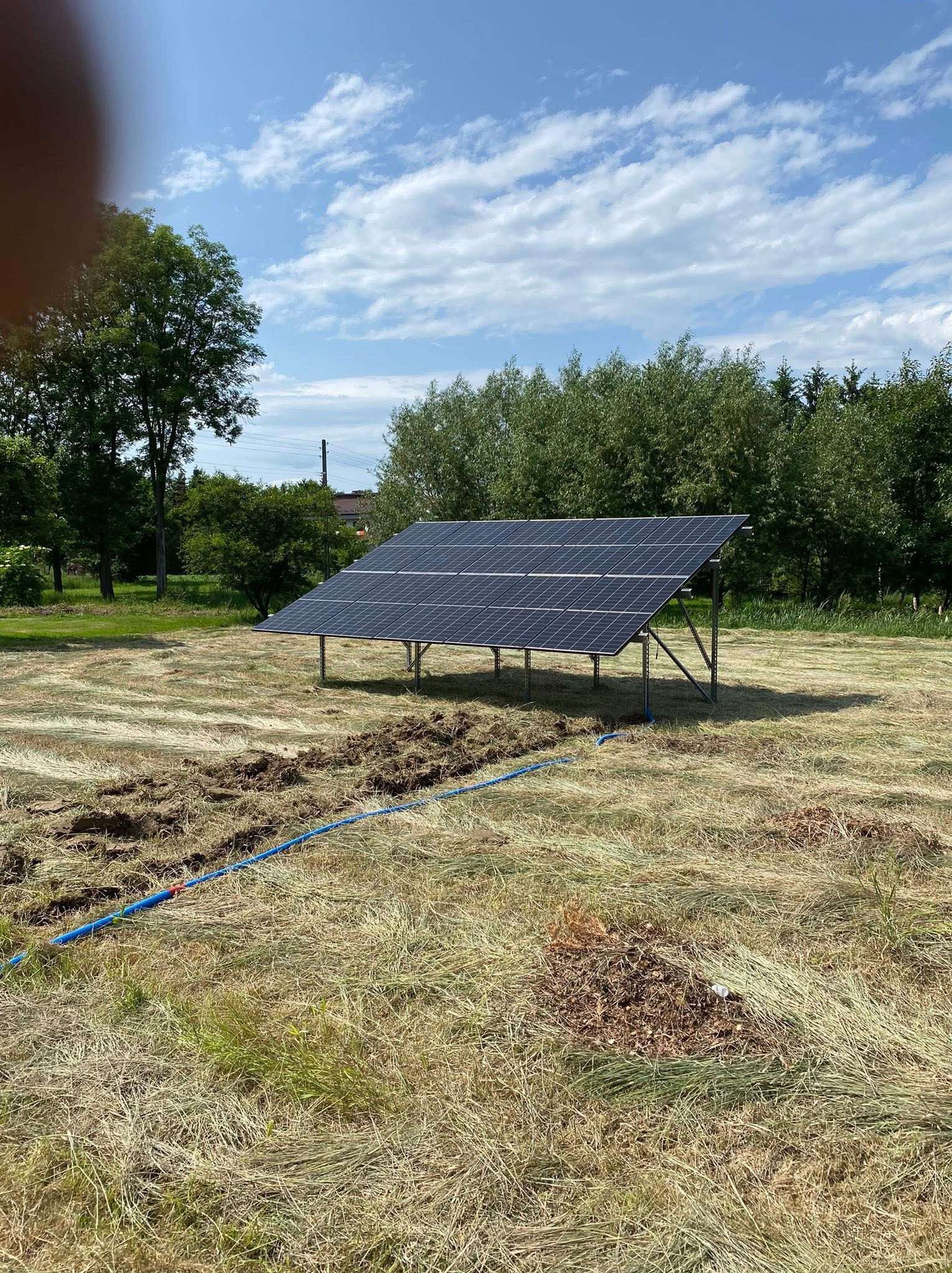 Panel fotowoltaiczny na łące, z niebieskim przewodem. Widoczne drzewa i błękitne niebo z chmurami. Montaż paneli na trawie.