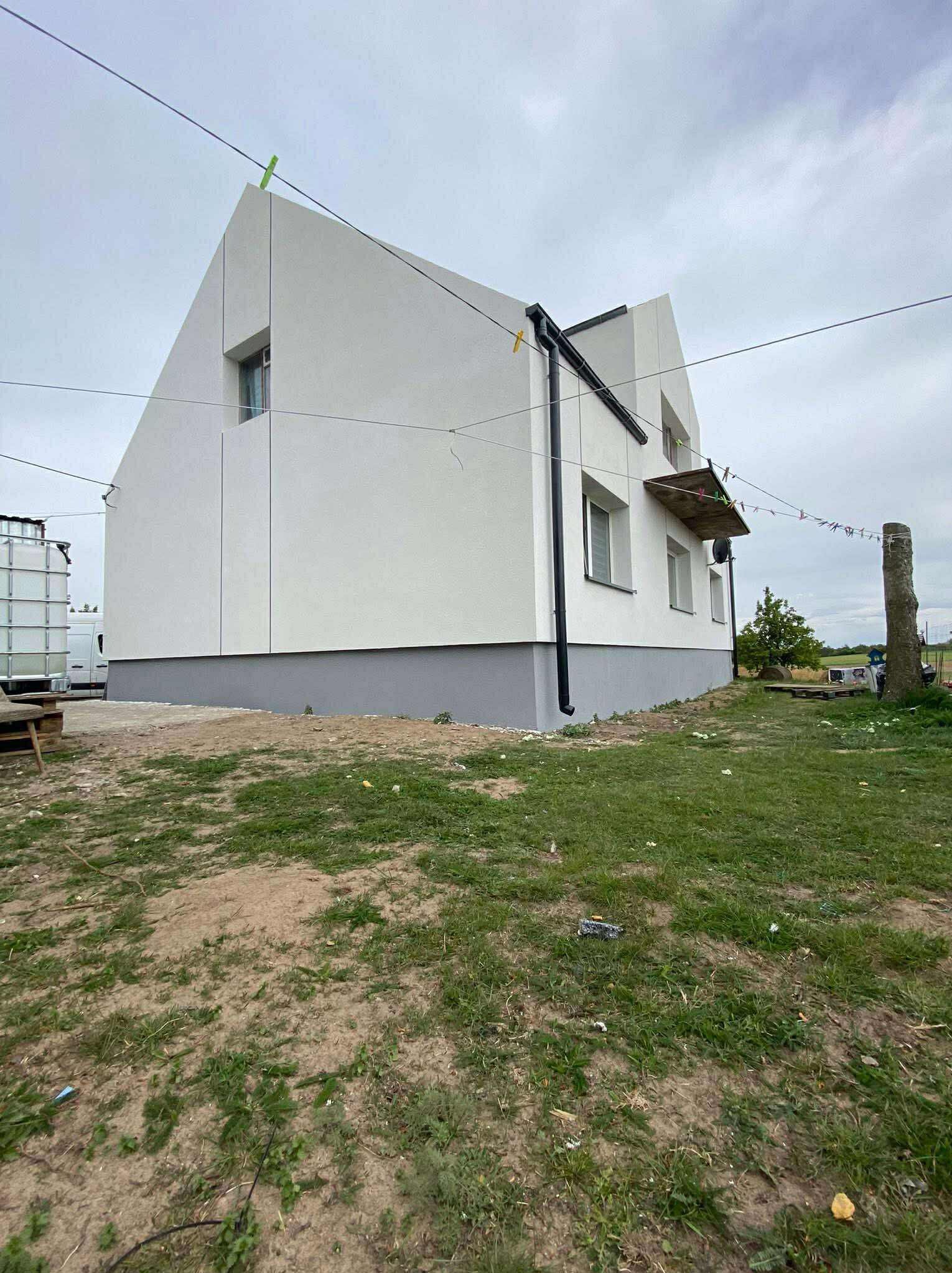 Nowoczesny dom z białą elewacją i szarym cokołem, widok z trawnika. Na ścianie widoczne rury spustowe. Suszące się pranie na zewnątrz.