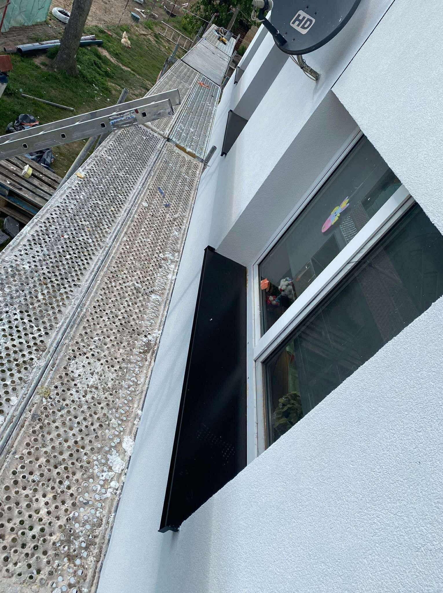 Świeżo otynkowana elewacja budynku w Krakowie, widoczna z rusztowania. Nowy tynk o grubej fakturze, okno z zabawkami w środku i antena satelitarna.