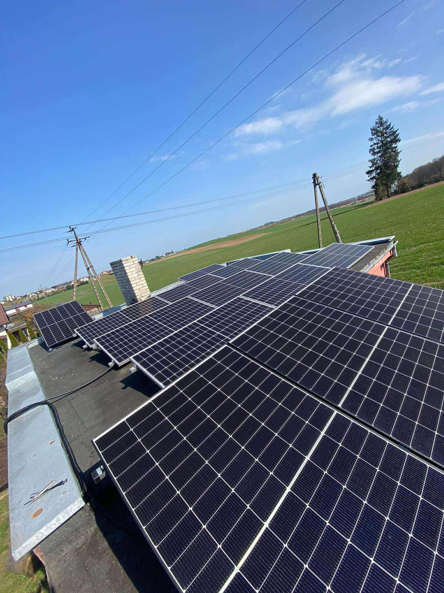 Panele fotowoltaiczne na dachu budynku z ceglanym kominem, na tle zielonego pola i błękitnego nieba z liniami energetycznymi.