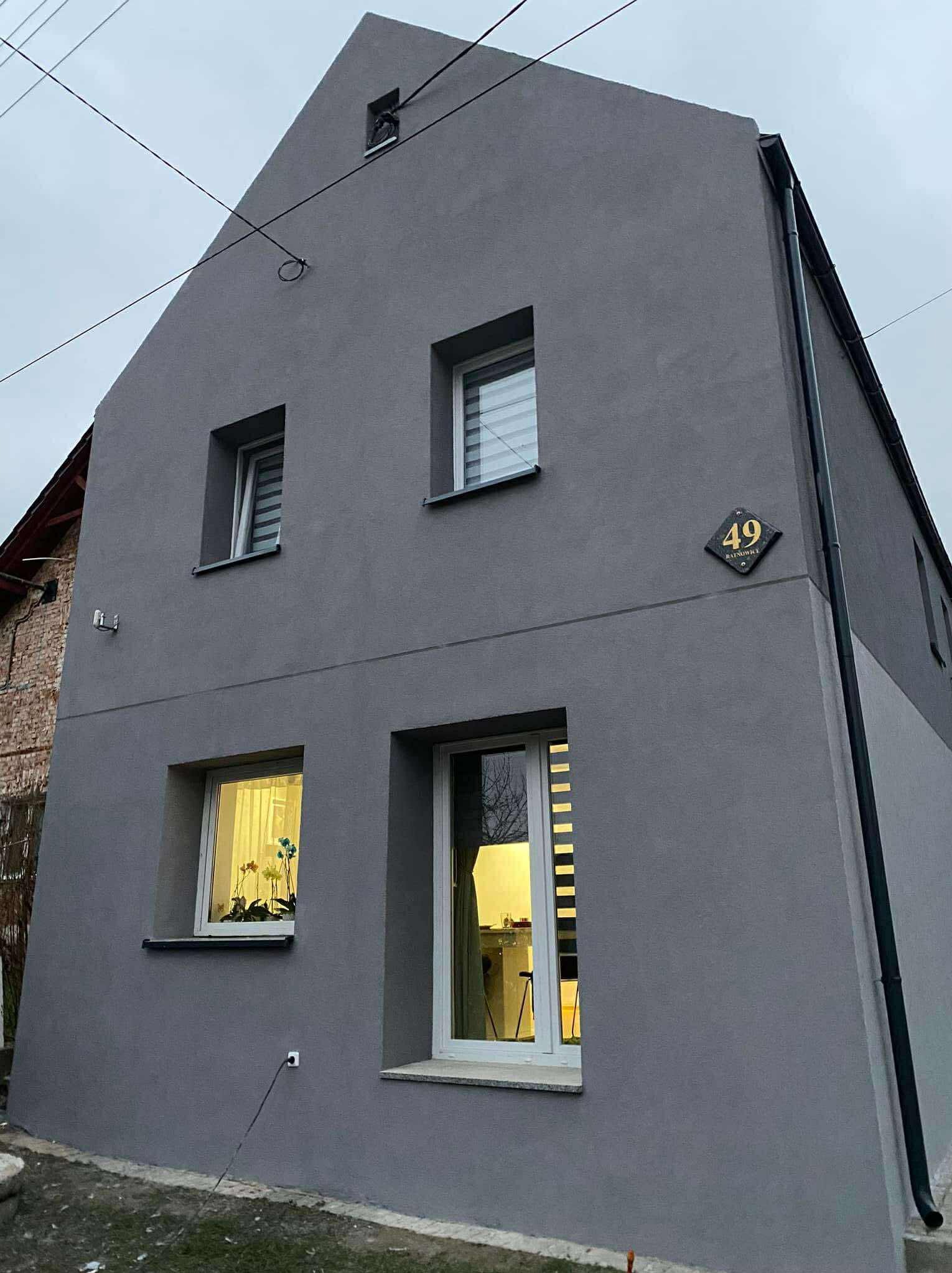 Szara elewacja domu z numerem 49, widok z dołu. Okna z żaluzjami, kwiaty w oknie. Nowoczesny styl, kontrast z ceglanym budynkiem obok.