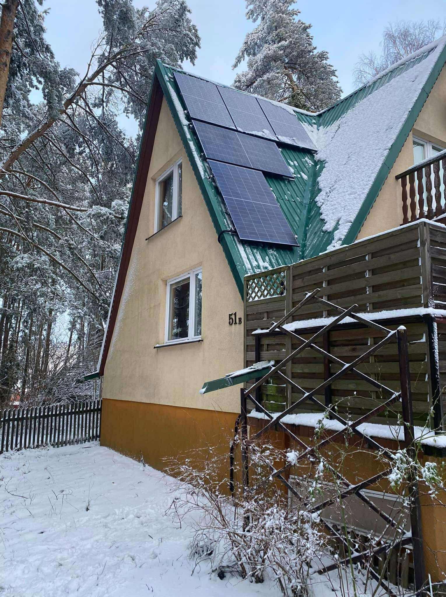 Dom z panelami fotowoltaicznymi na zielonym dachu pokrytym śniegiem. Zimowy krajobraz, drzewa oszronione. Widok z poziomu gruntu.