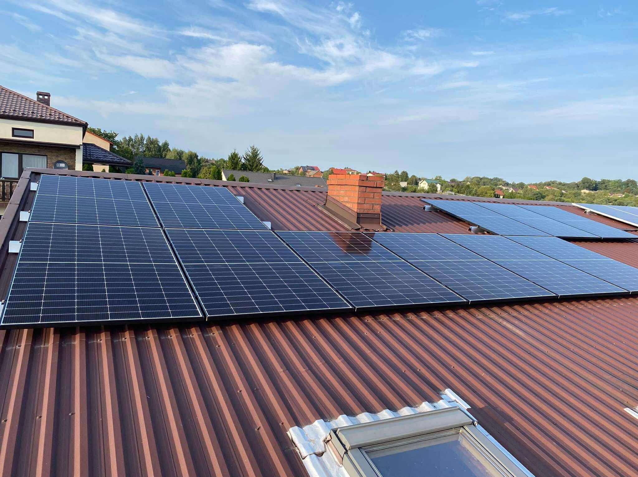 Panele fotowoltaiczne na brązowym dachu z blachy falistej, widok z góry, fragment komina z cegły w tle, błękitne niebo z chmurami.