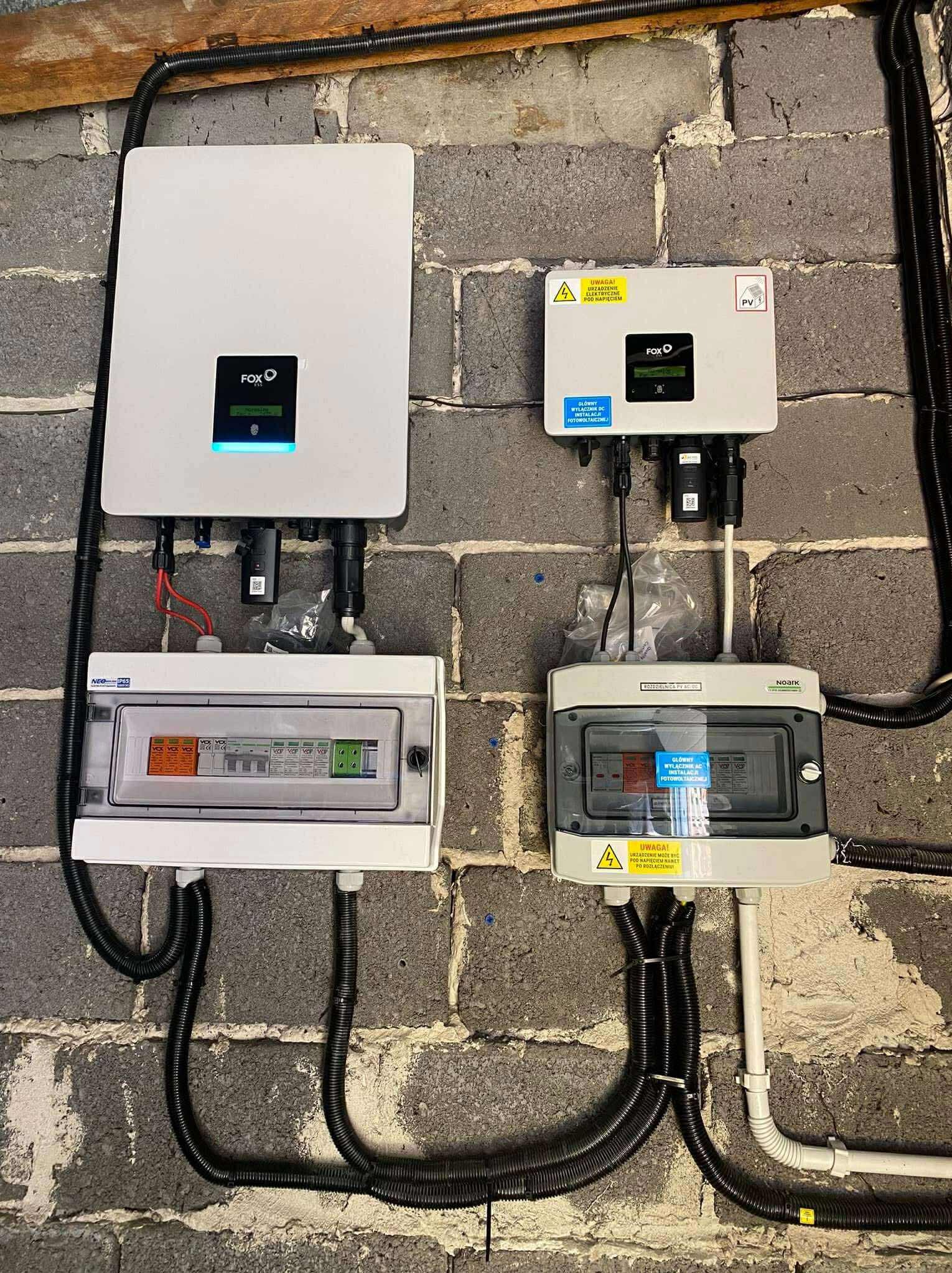 Dwa falowniki FOXO i rozdzielnice AC/DC zamontowane na szarej, murowanej ścianie. Widoczne kable w peszlach. Instalacja fotowoltaiczna wewnątrz budynku.