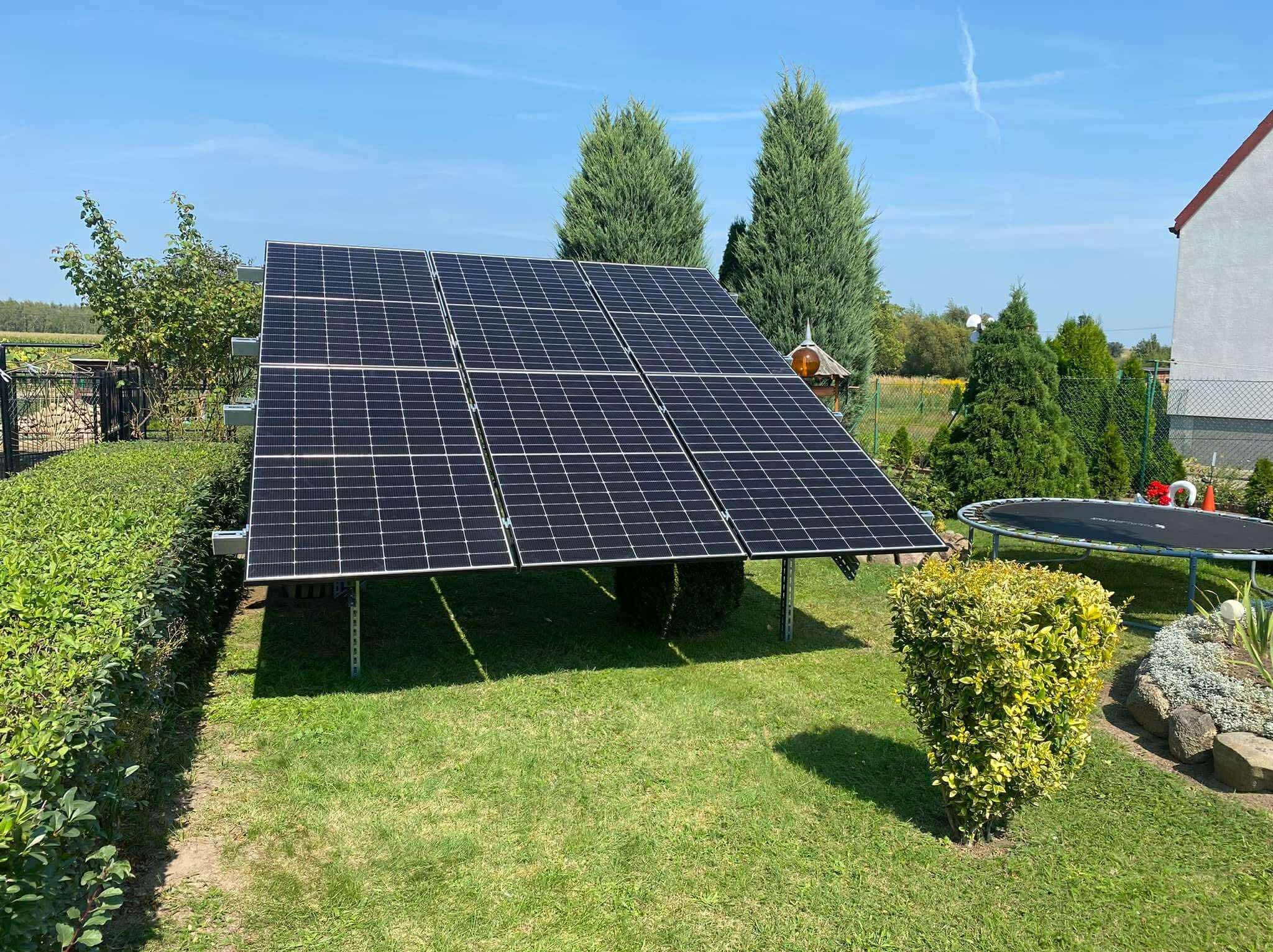 Panele fotowoltaiczne na trawniku, widok z boku. W tle zielone drzewa i fragment domu. Instalacja na metalowej konstrukcji, w otoczeniu krzewów.