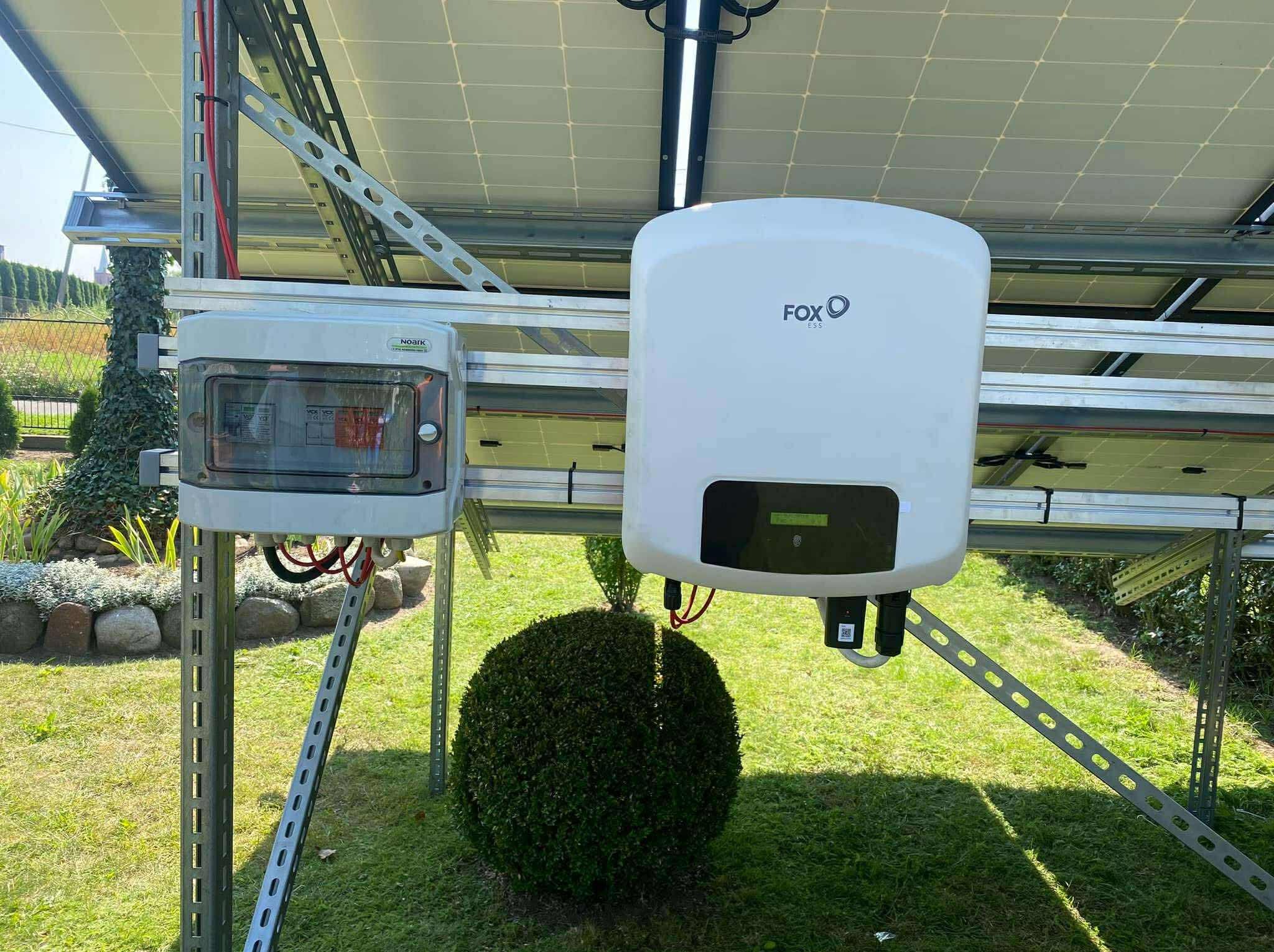 Instalacja fotowoltaiczna: biały inwerter Fox ESS i szara skrzynka Noark na metalowej konstrukcji, panele słoneczne, trawnik i krzew. Widok z poziomu gruntu.