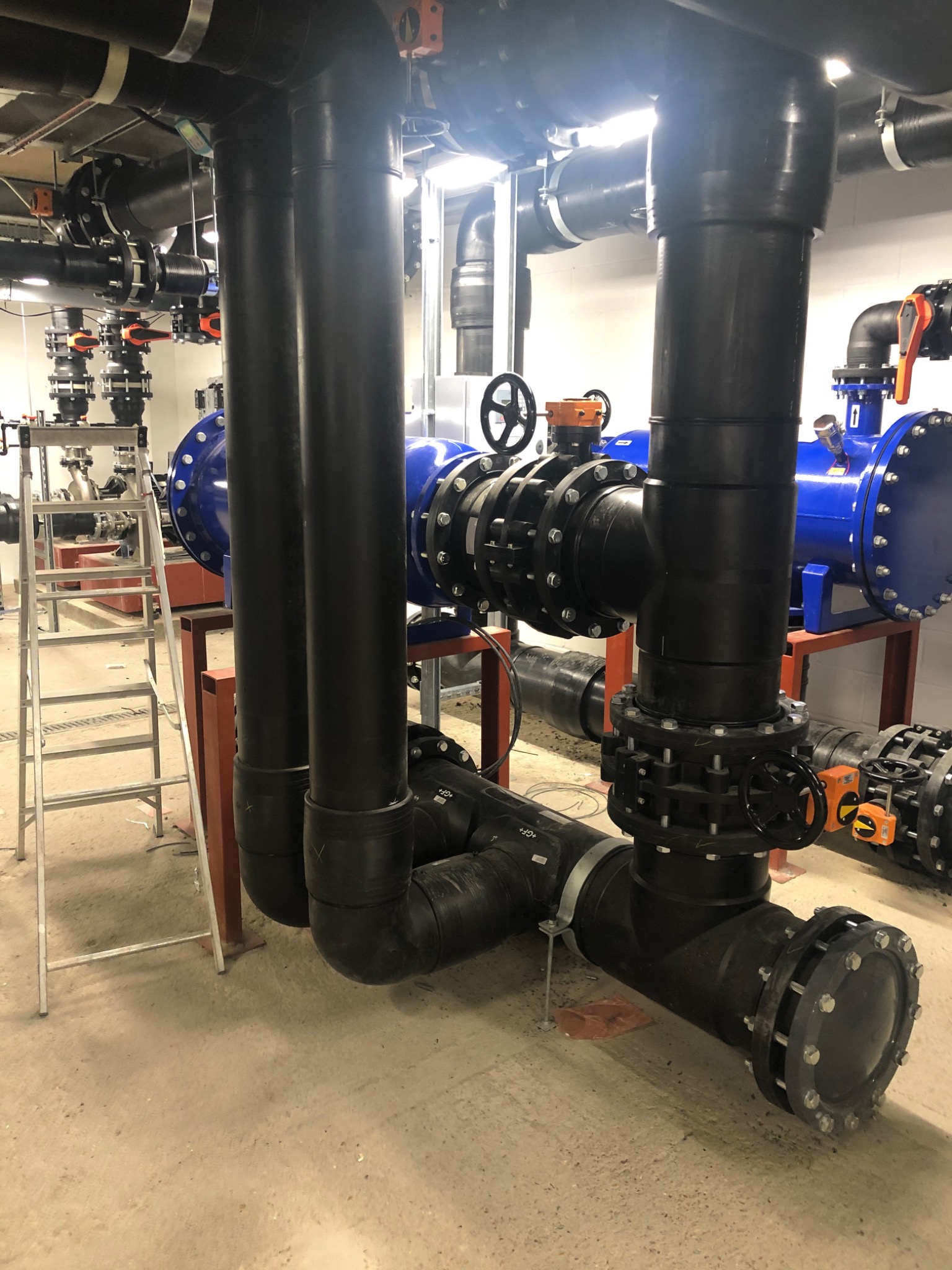 Złożona instalacja hydrauliczna z rurami z tworzywa sztucznego, zaworami i armaturą. Widoczna drabina i elementy konstrukcyjne. Wykończona instalacja w pomieszczeniu technicznym.