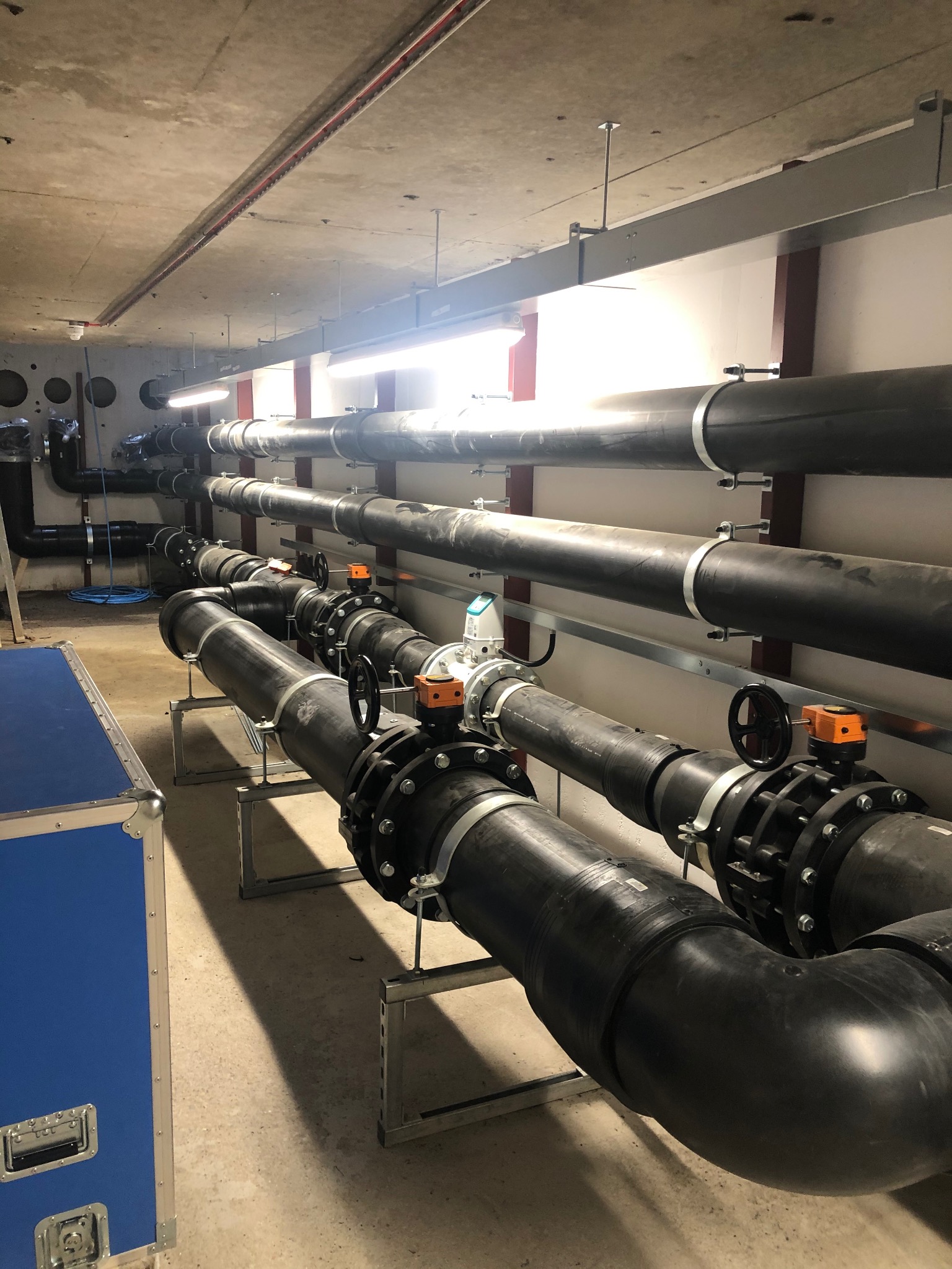 Czarne rury instalacji przemysłowej z zaworami, zamocowane na wspornikach pod sufitem w surowym wnętrzu. Widoczne łączenia i mocowania.