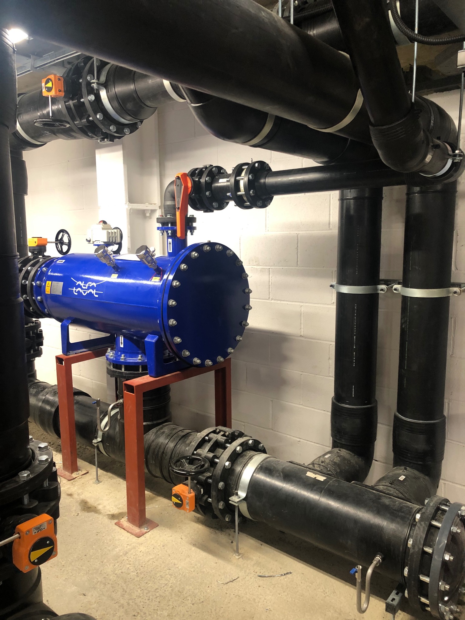 Kompleksowa instalacja rur przemysłowych z filtrem ciśnieniowym Alfa Laval, z licznymi złączami kołnierzowymi i zaworami, zamontowana w pomieszczeniu technicznym.