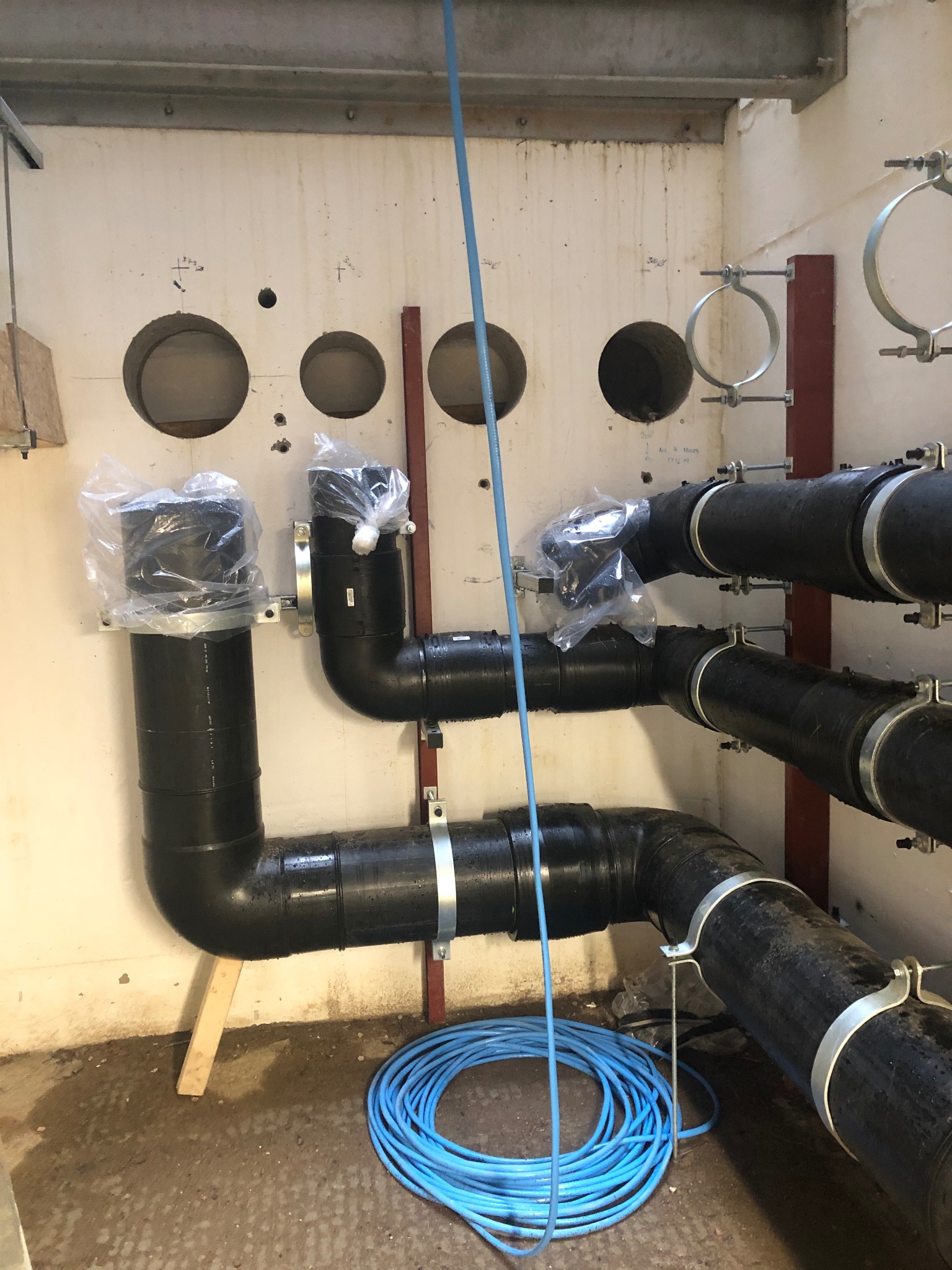 Czarne rury kanalizacyjne z obejmami mocowane do ściany, zabezpieczone folią, niebieski kabel na podłodze, widoczne otwory w ścianie.