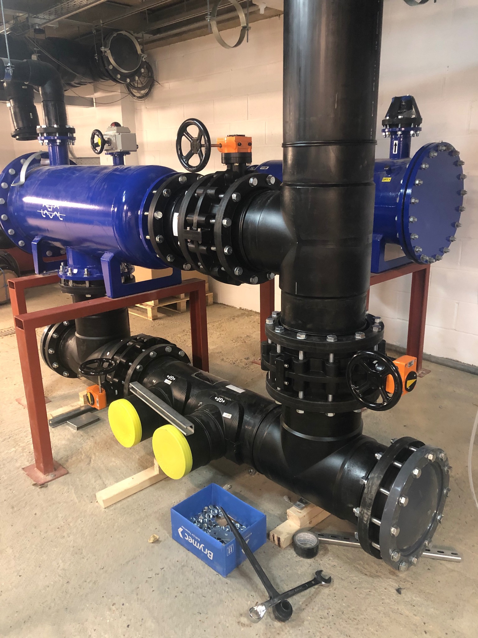 Złożona instalacja rur z zaworami i filtrami, wsparta na stalowej konstrukcji. Widoczne narzędzia i elementy montażowe, sugerujące etap prac instalacyjnych.