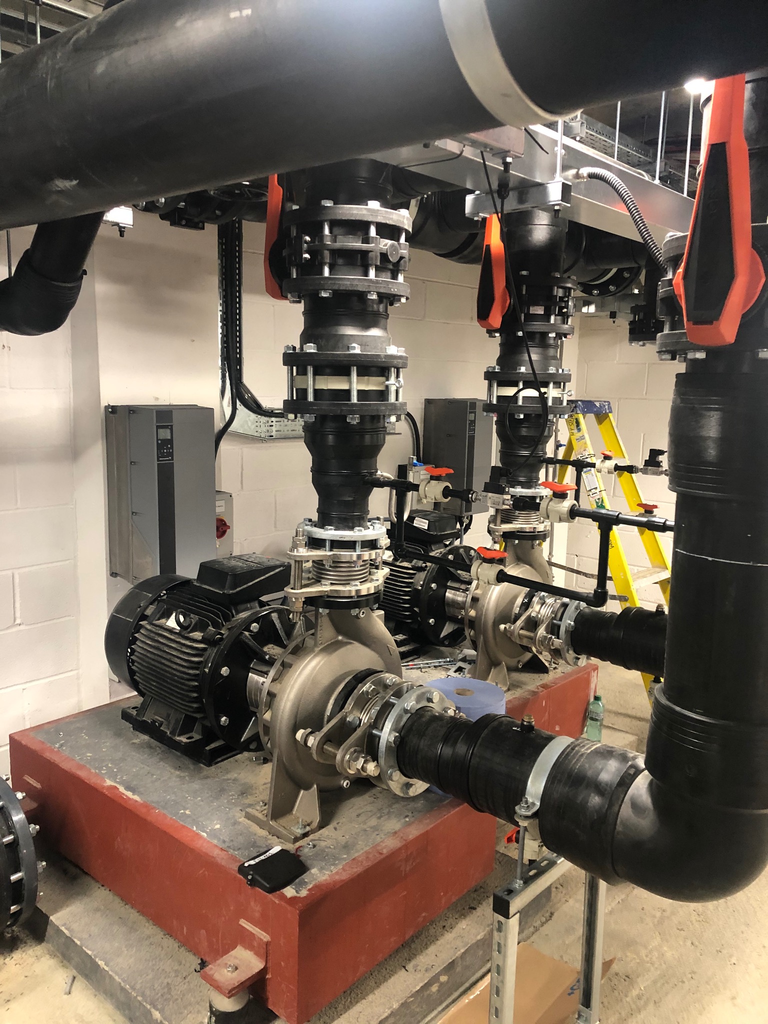 Złożona instalacja pomp i rur w kotłowni w Kostrzynie. Widoczne czarne rury, pompy na czerwonej podstawie i drabina. Elementy instalacji połączone śrubami.