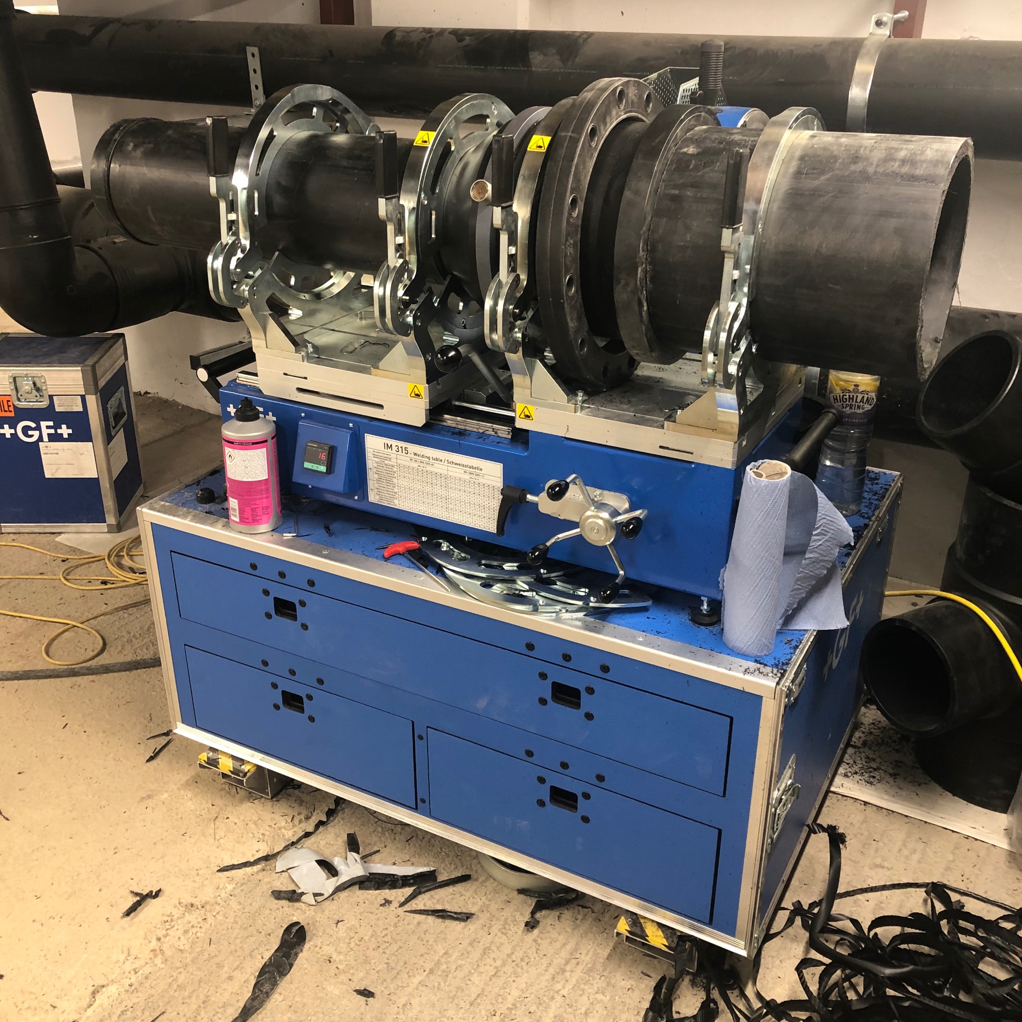 Zgrzewarka doczołowa IM 315 do rur z tworzyw sztucznych, z zamocowanymi rurami, na niebieskim wózku narzędziowym w pomieszczeniu. Widoczne akcesoria i odpady z obróbki.